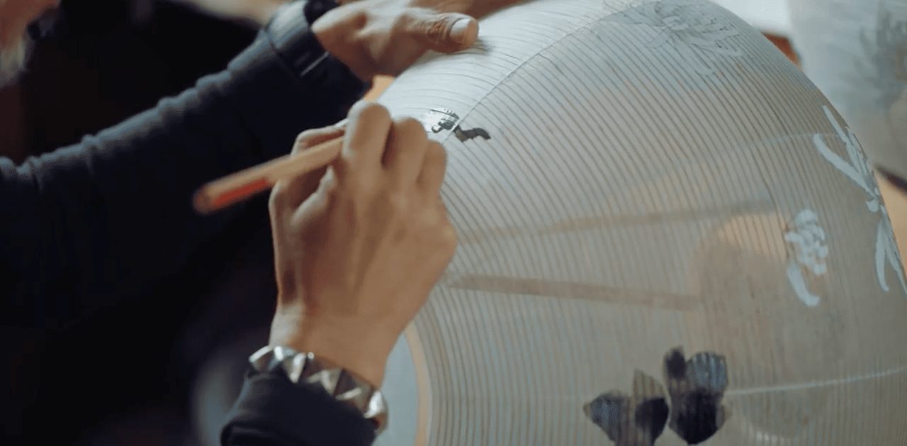 Hands painting a small dark motif onto the ribbed paper of a large white lantern with a fine brush.