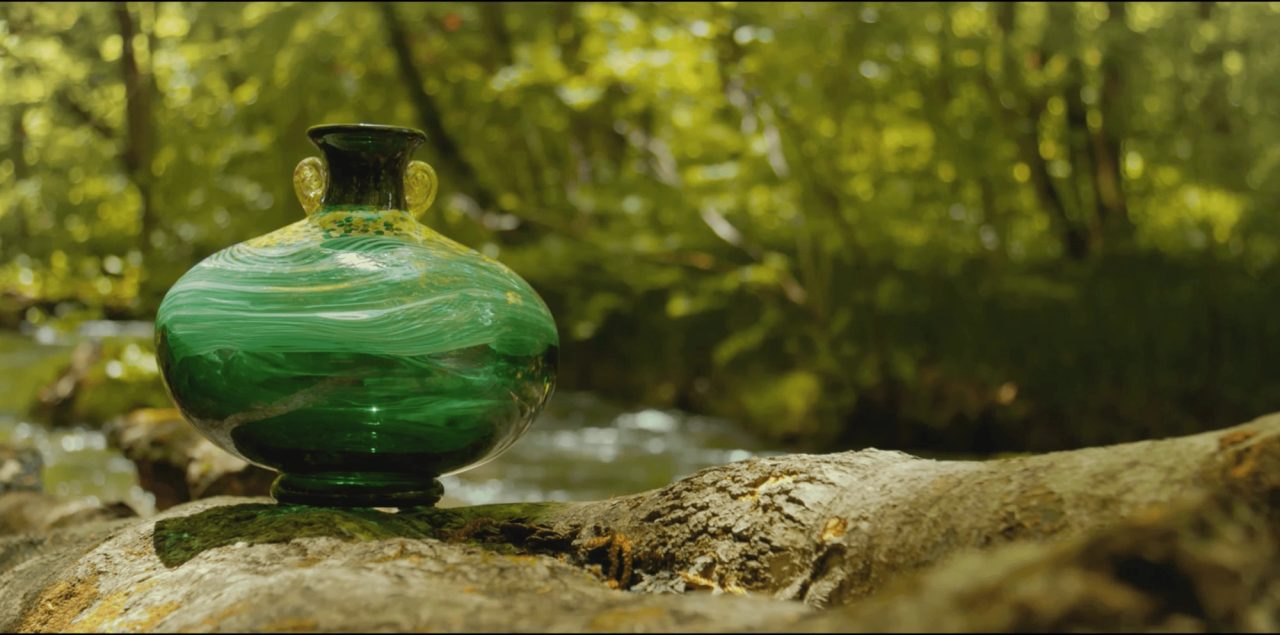 A round green glass vessel with swirled bands and two small handles, standing on a mossy rock beside a stream in woodland.