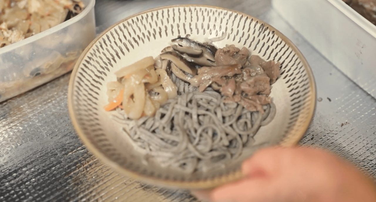 A hand holding a glazed bowl chipped all over with fine tool marks around the rim, filled with grey noodles, mushrooms and vegetables.