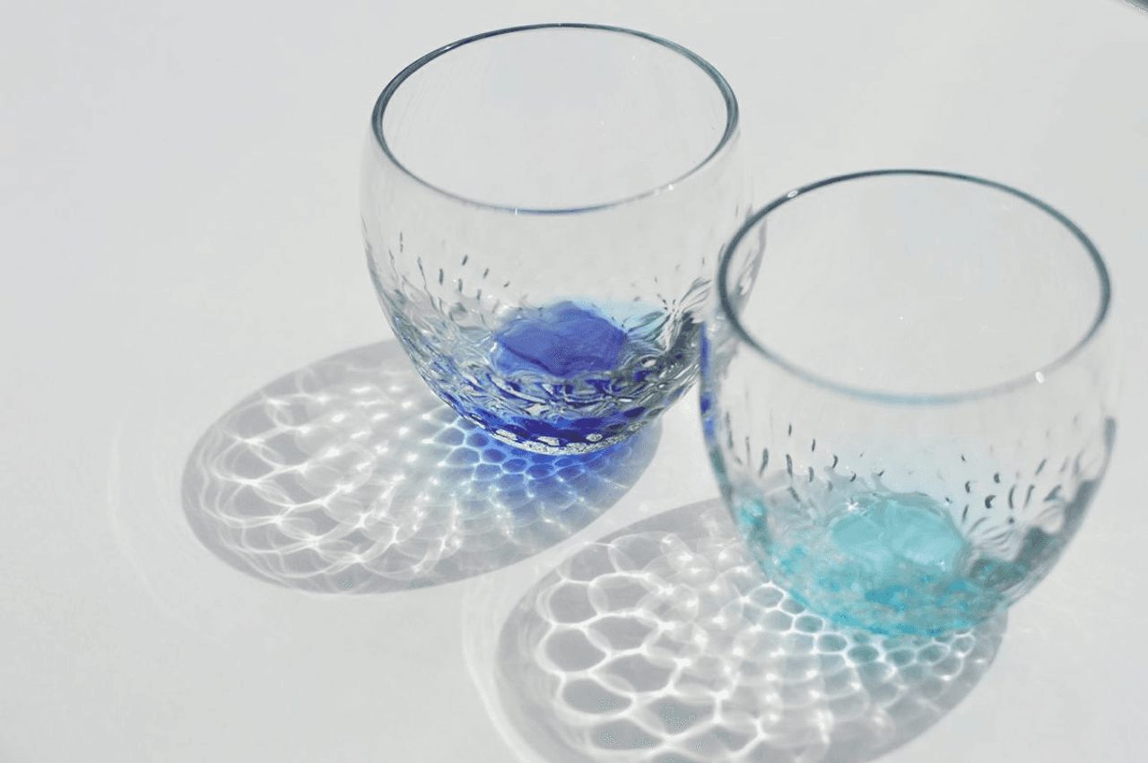 Two blown glass tumblers on a white surface, one with a cobalt base and one turquoise, casting dappled shadows through their bubbled walls.