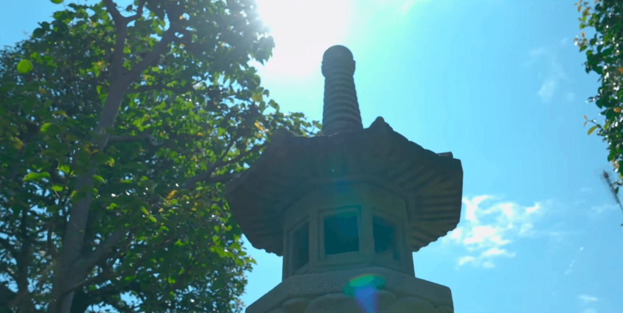 A carved granite lantern photographed from below against a bright blue sky, sunlight flaring behind its tiered stone roof and finial.