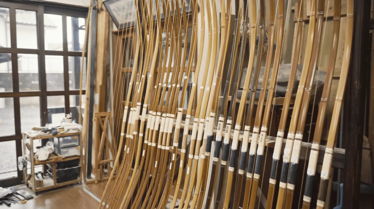 Dozens of tall bamboo bows standing in rows against a workshop wall, their grips bound in white and black cord, daylight from windows behind.