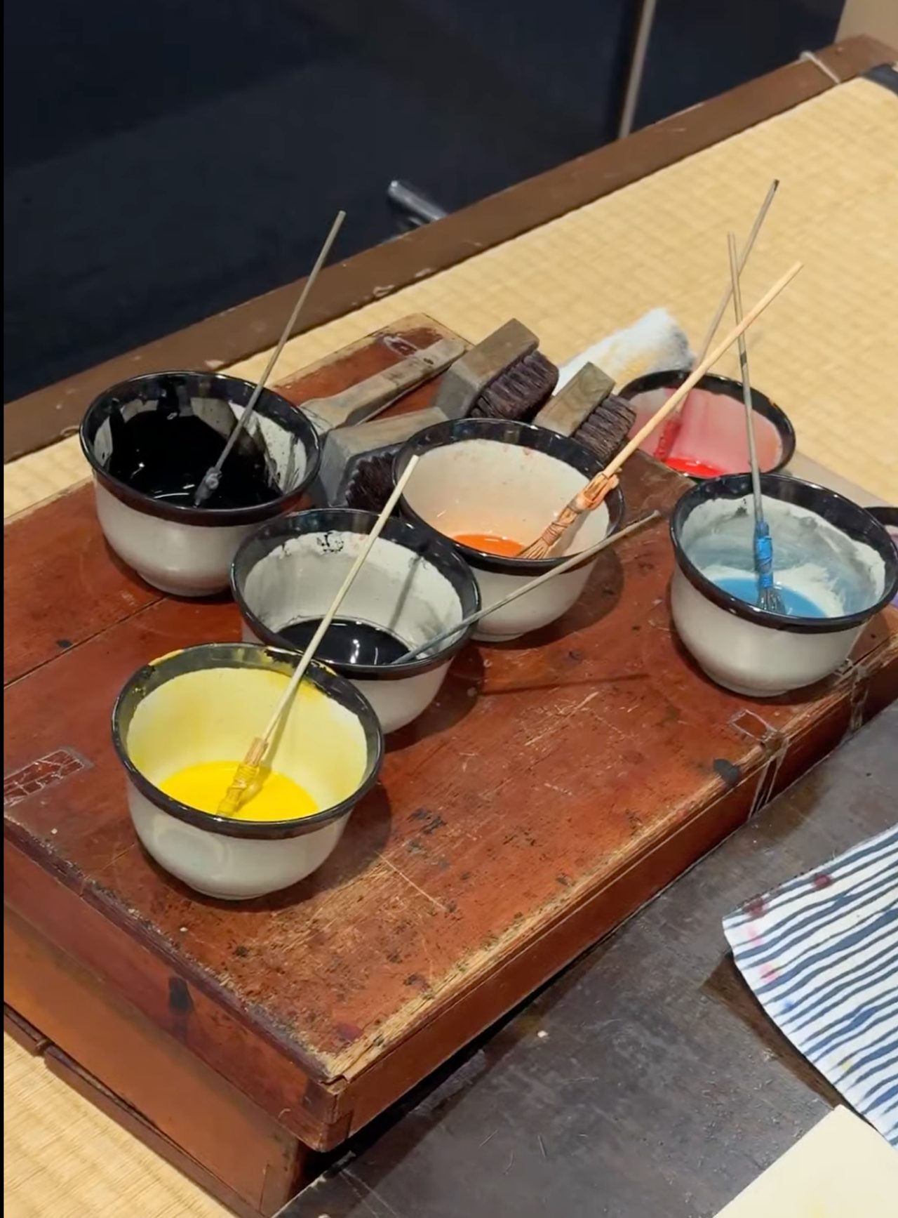 Bowls of woodblock printing pigment with brushes resting in them, on a worn wooden board.