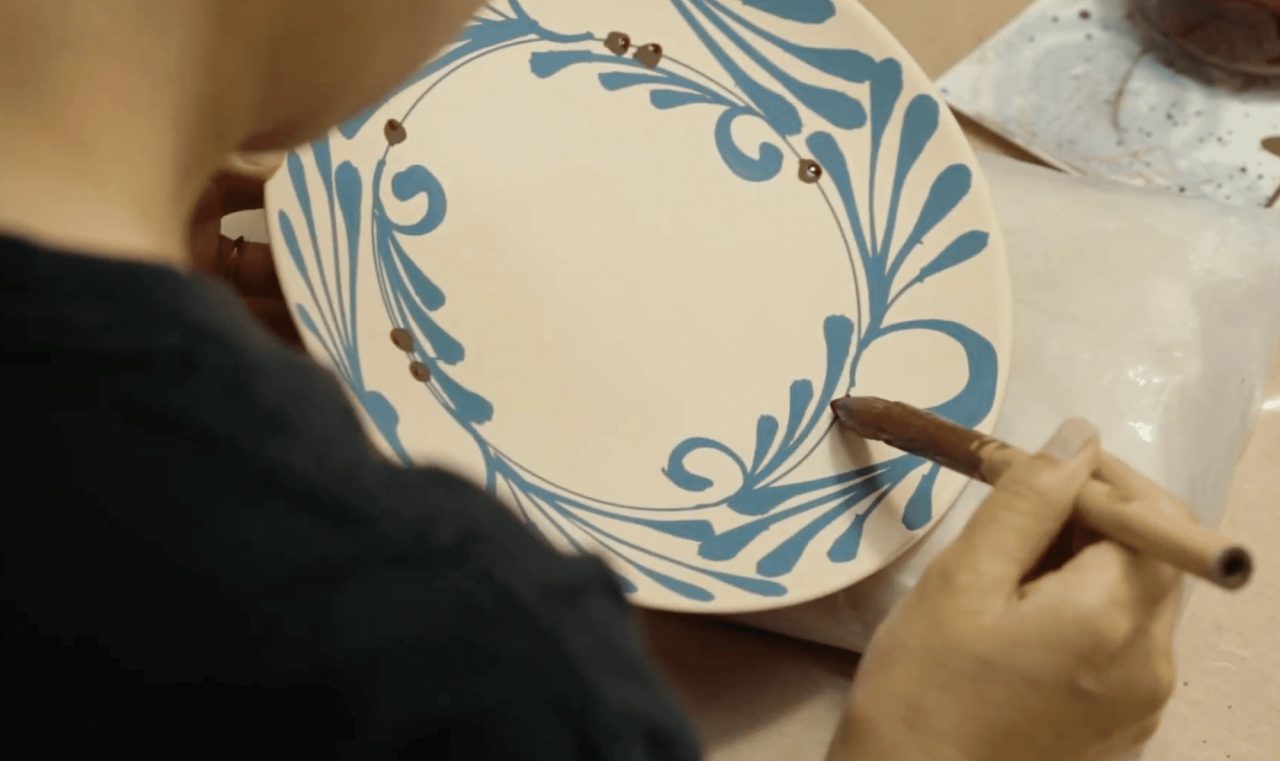 Seen over a shoulder, a hand painting a blue scrolling border onto a white porcelain plate with a slim brush.