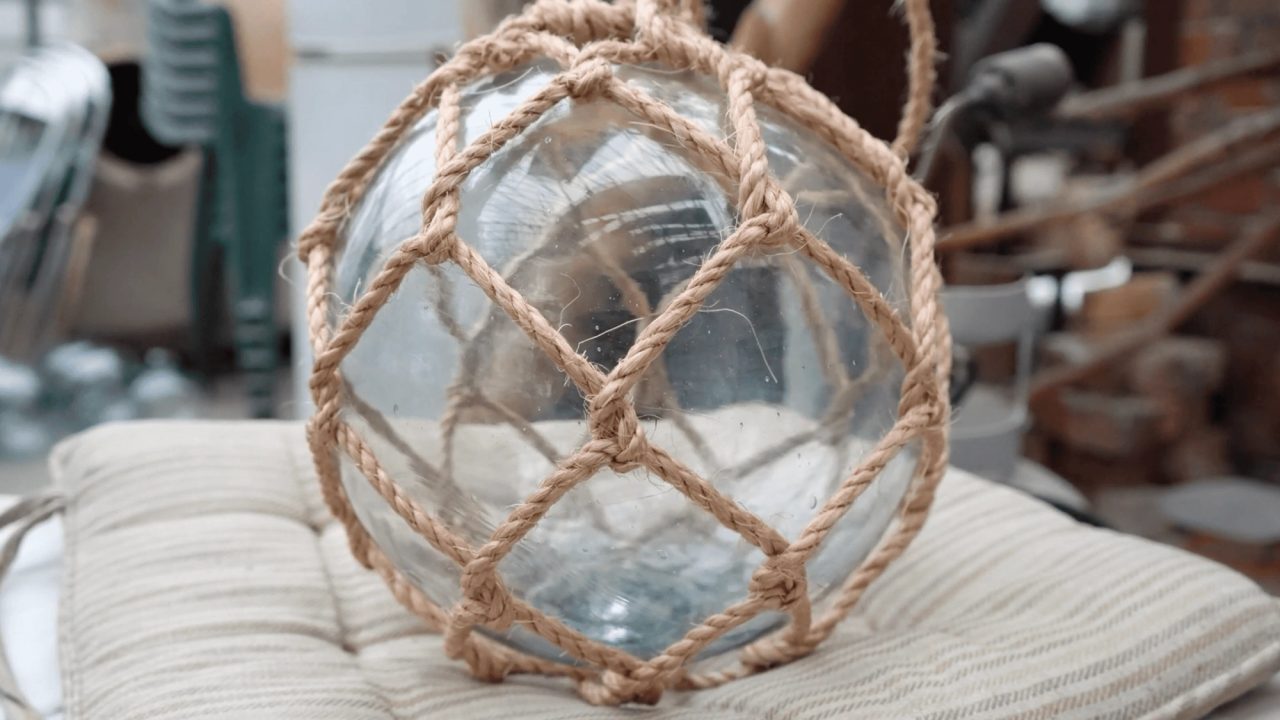 A clear glass fishing float wrapped in a knotted rope net, resting on a padded cushion in a workshop.