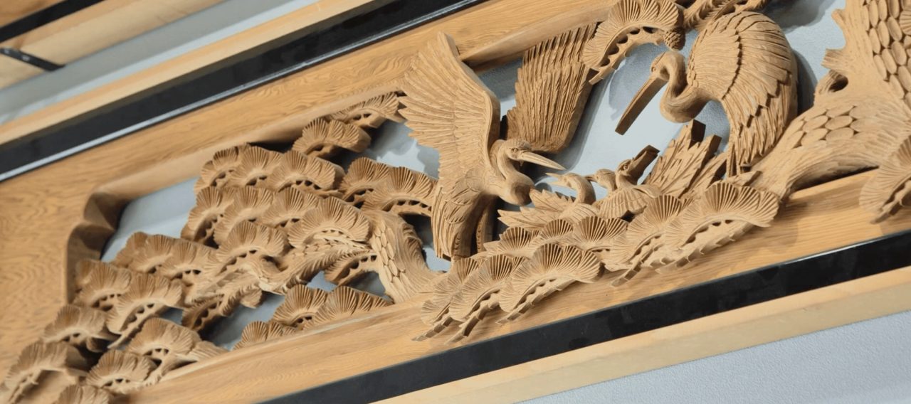 A carved wooden transom panel set above a doorway, showing two cranes among pine branches cut in deep relief.
