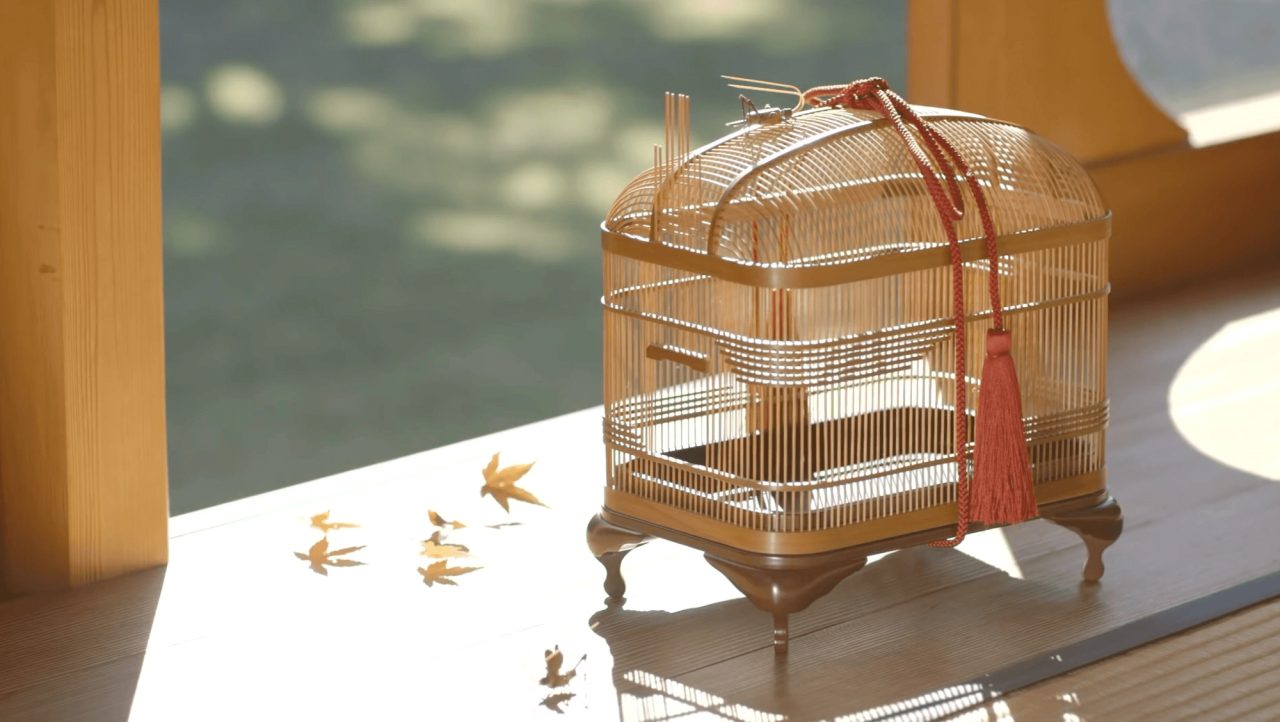 A domed birdcage of very fine bamboo rods on a footed stand, hung with a red tassel, standing on a veranda beside scattered maple leaves.