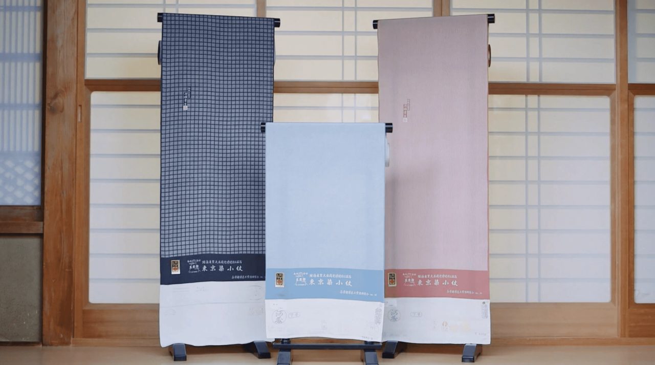 Three bolts of finely patterned cloth hung on stands before paper screens, in dark check, pale blue and dusty pink.
