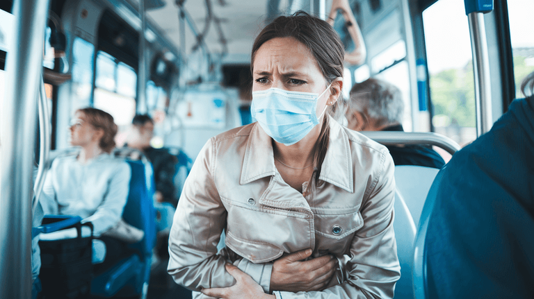 A woman sitting on a bus wearing a medical mask looks worried and holds her stomach