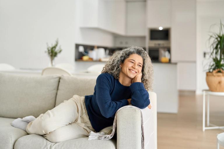 Smiling woman relaxing on a sofa in a bright, cozy home, expressing a sense of calm, reassurance, and emotional ease.
