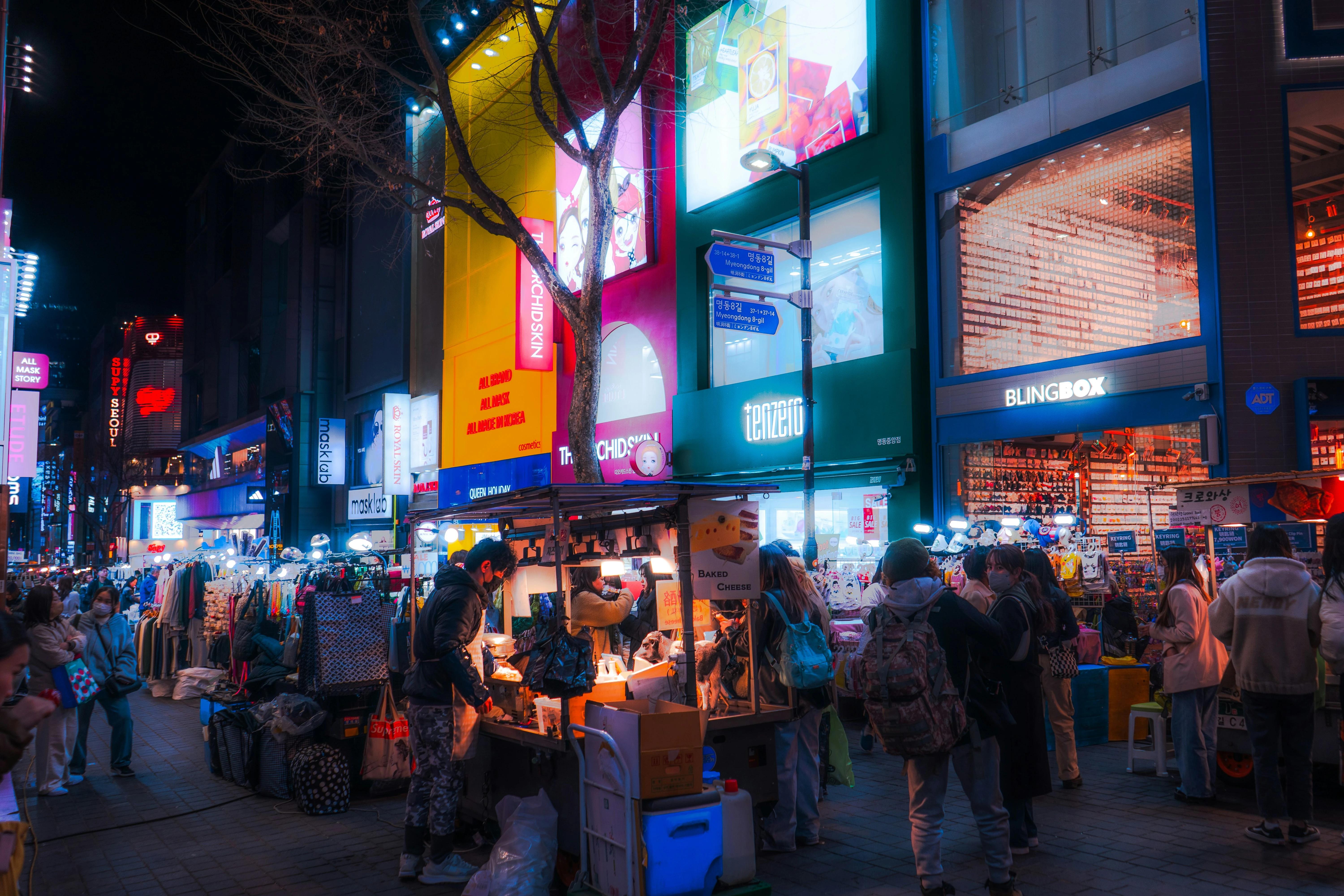 Busy Myeongdong shopping street at night with neon-lit skincare shops, street food stalls, and crowds of shoppers