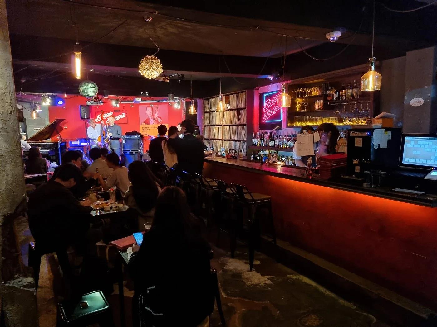 Live jazz performance at Boogie Woogie bar in Itaewon with saxophonist on stage, vinyl-lined shelves, and neon signage behind the bar