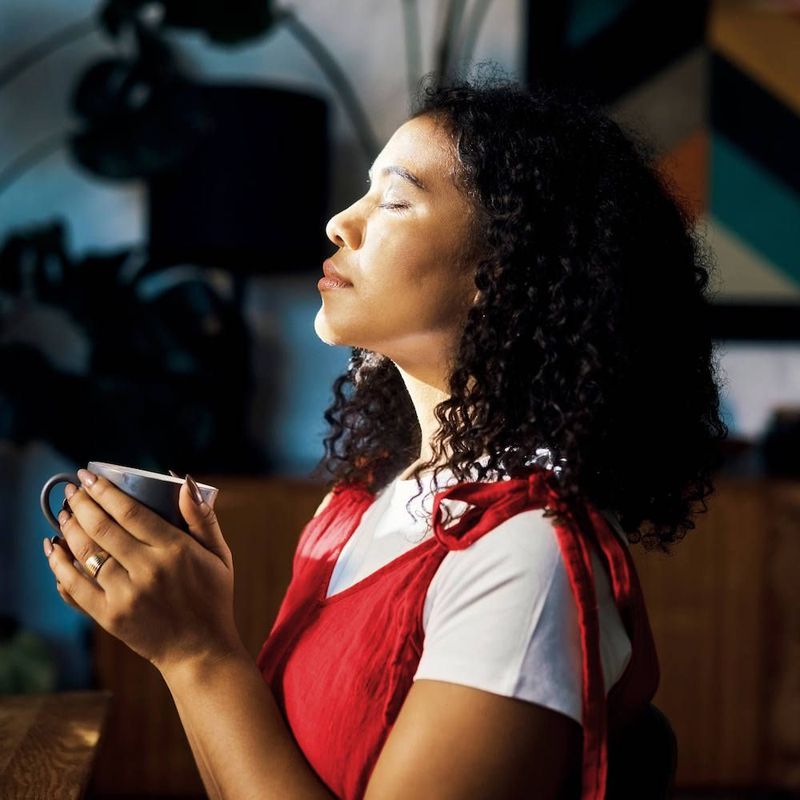 Photo of a woman next to her living room window, standing with her eyes closed and her face in the sunlight