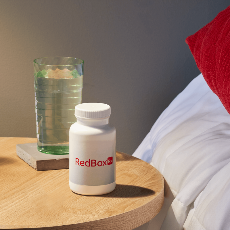 White prescription pill bottle labeled “RedBox Rx” on a bedside table next to a glass of water.