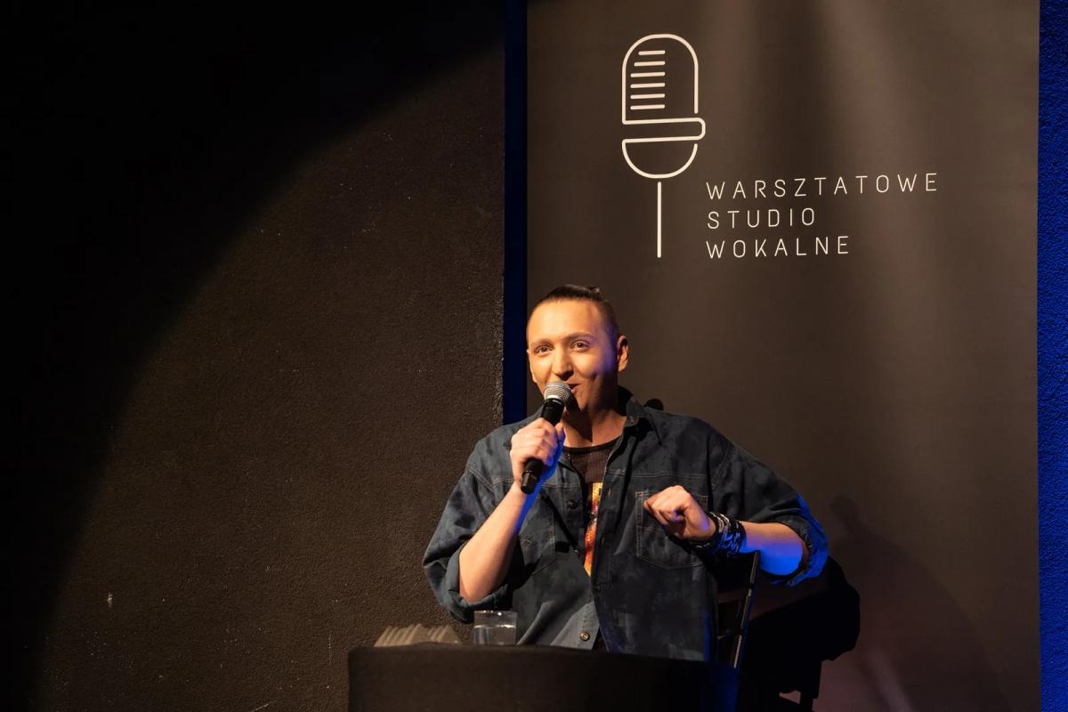 Prowadzący mówi do mikrofonu obok planszy Warsztatowego Studia Wokalnego.