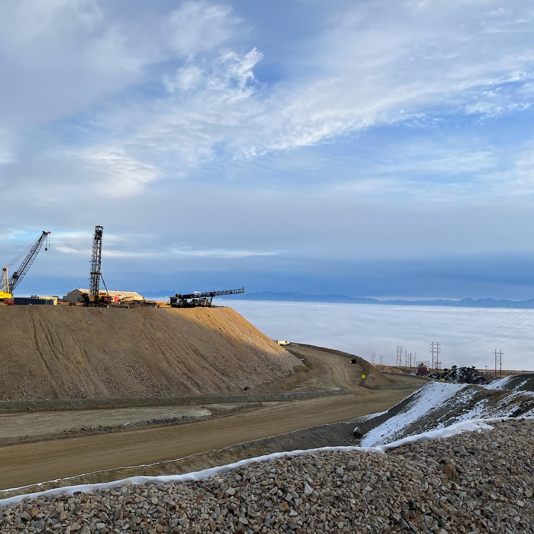 Cranes and drilling equipment operate at a snowy mountain mine above a cloud-filled valley.