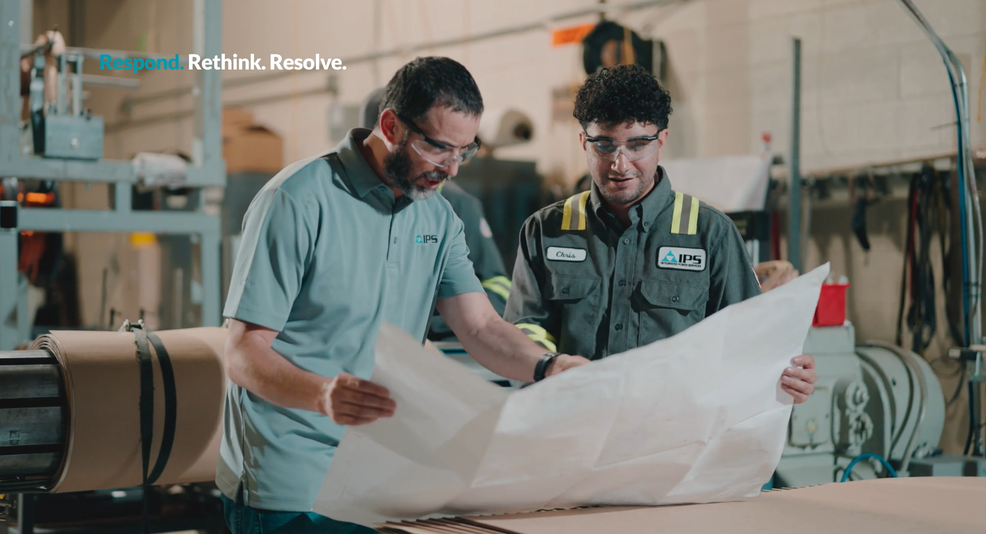 Two men wearing safety glasses review blueprints in an industrial setting.