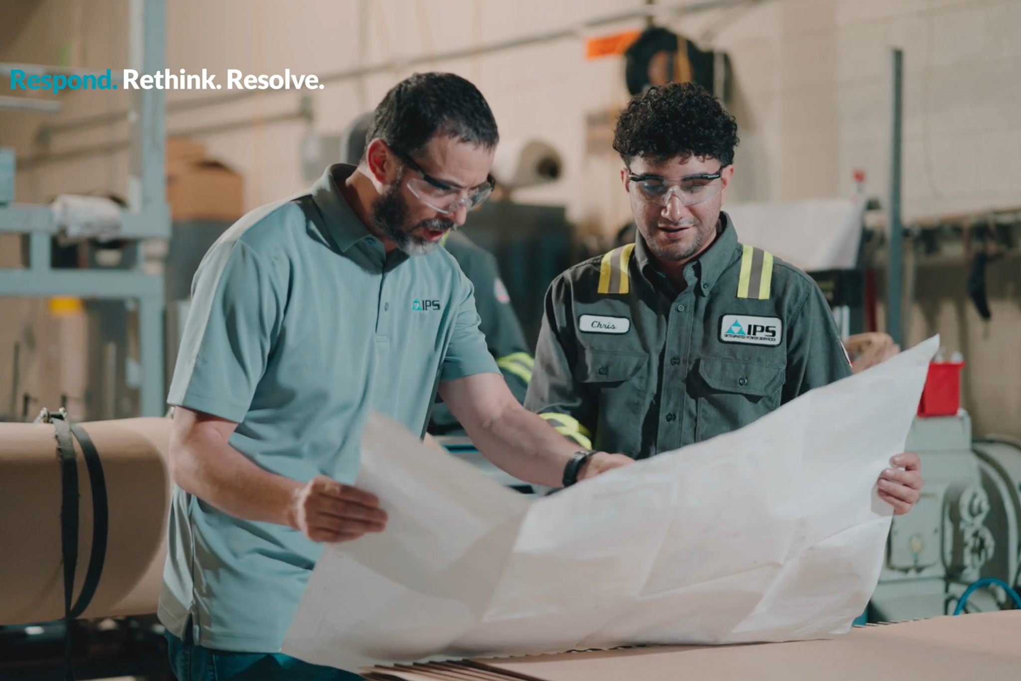 Two men wearing safety glasses review blueprints in an industrial setting.