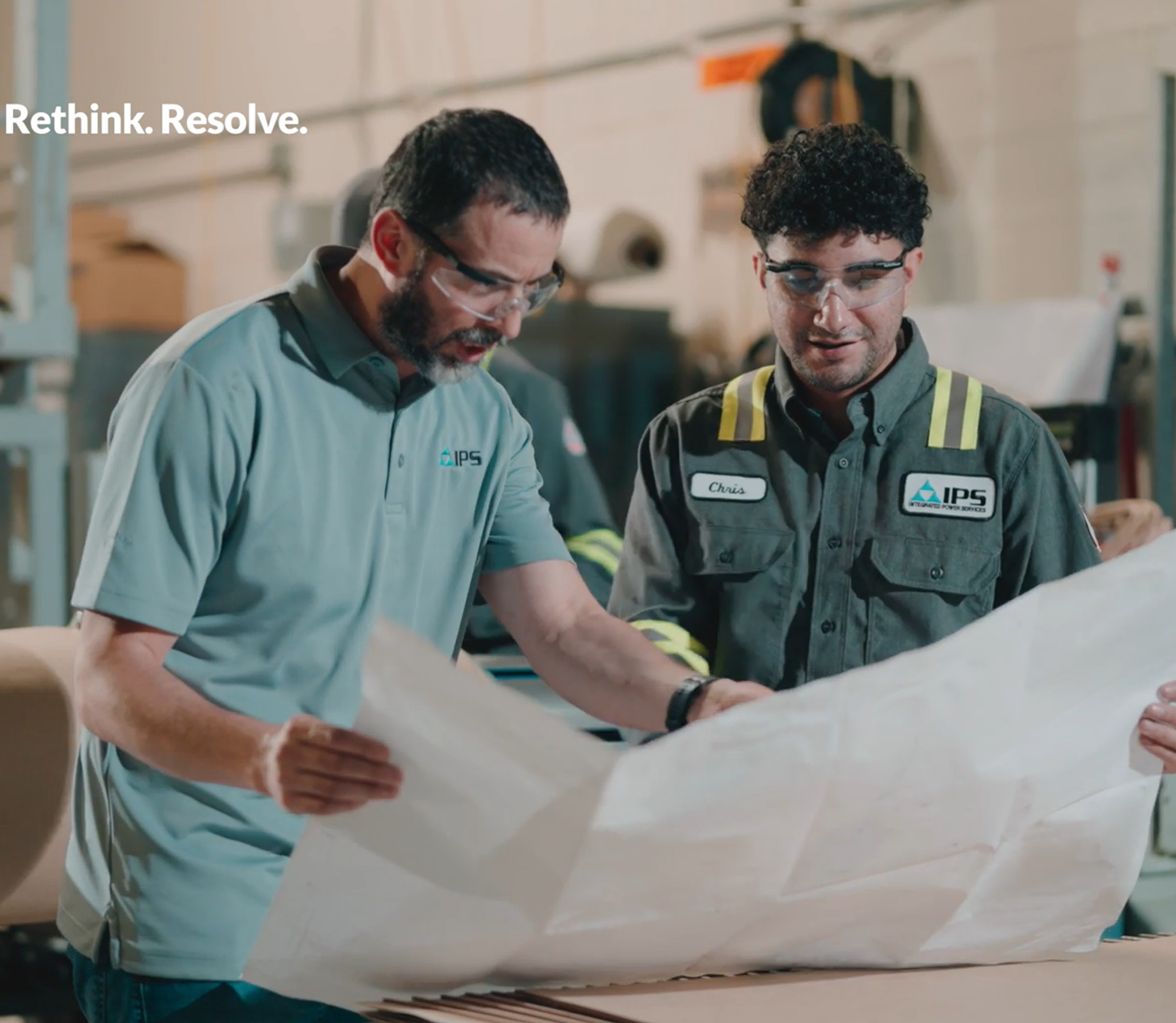 Two men wearing safety glasses review blueprints in an industrial setting.