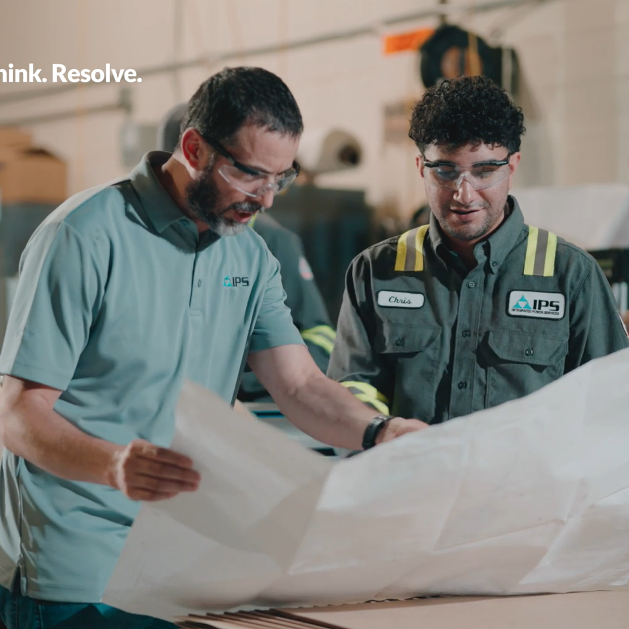 Two men wearing safety glasses review blueprints in an industrial setting.