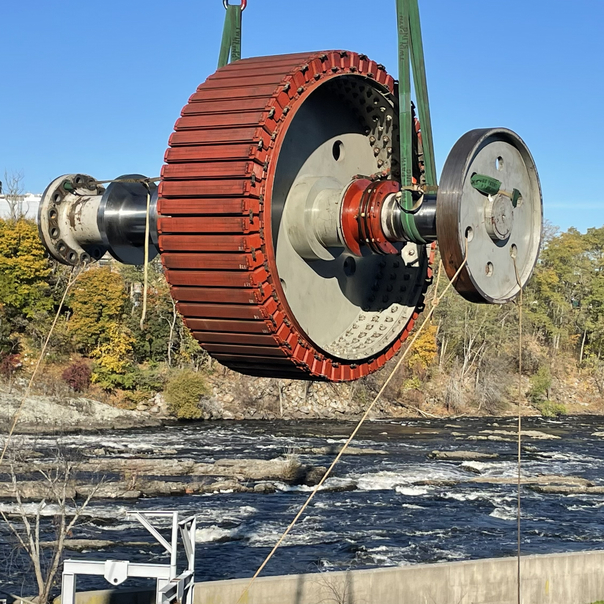 Crane lifts a large hydroelectric generator rotor above a river during maintenance.