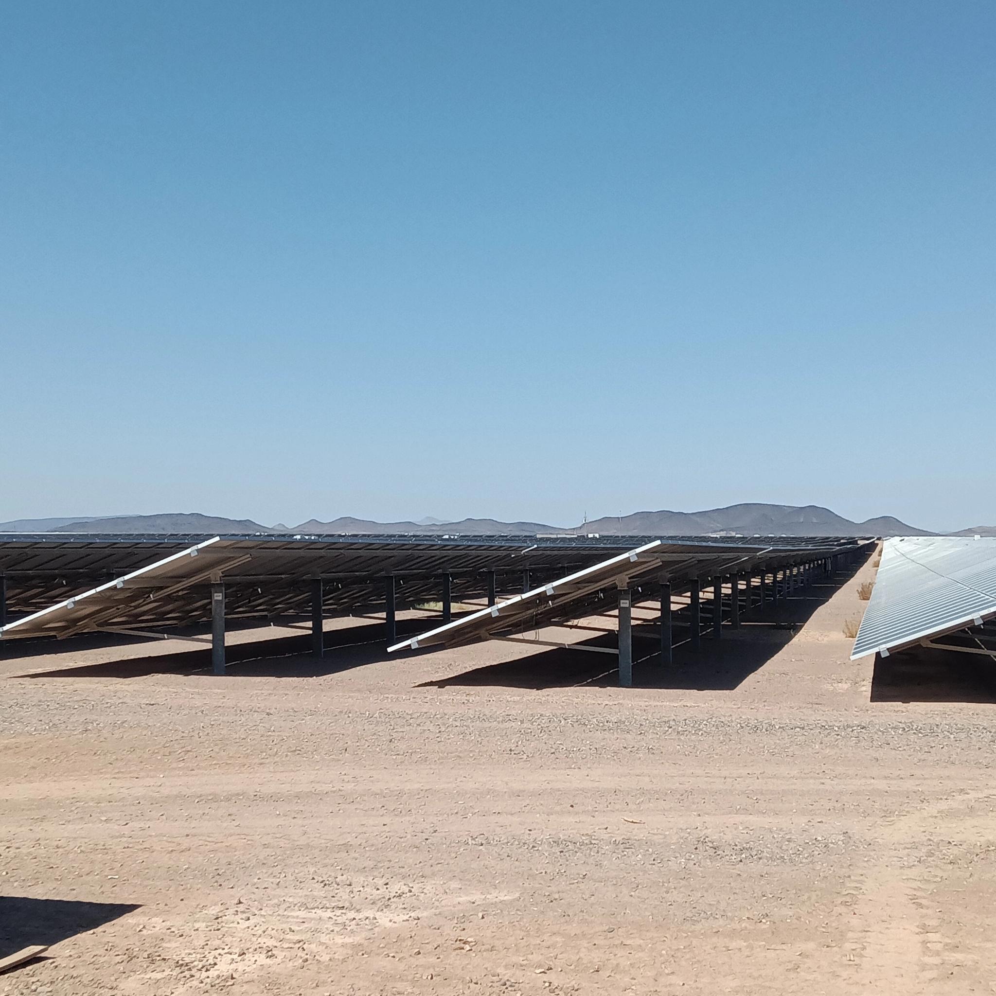 Rows of solar panels installed in a desert landscape with mountains in the distance.