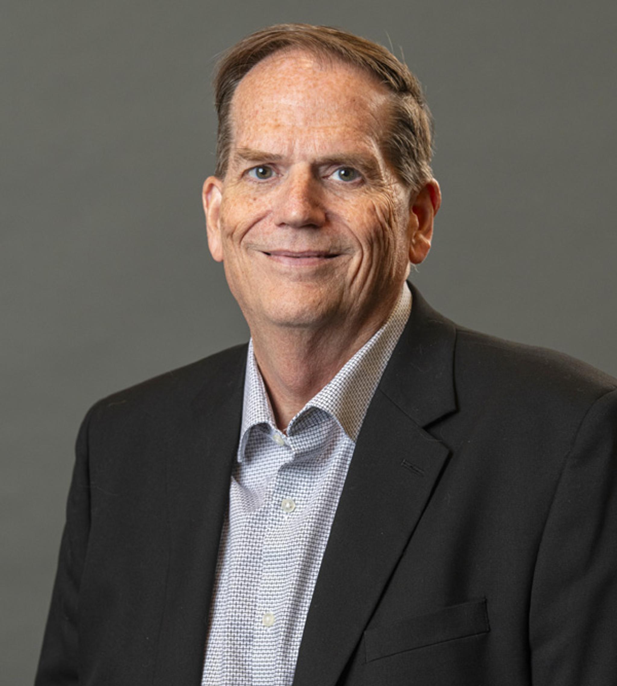 Professional headshot of a smiling man in a dark suit jacket against a gray background.