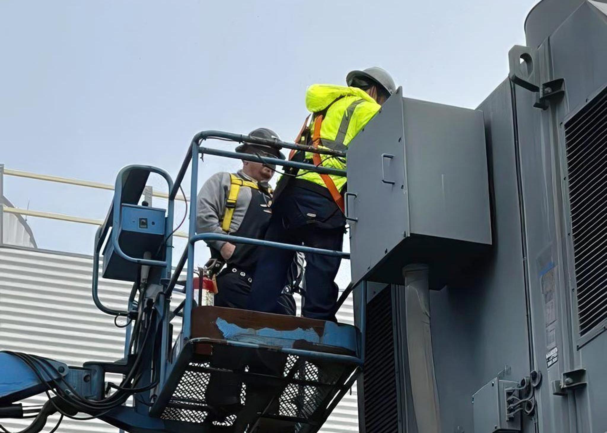 Technicians in safety harnesses inspect an electrical equipment cabinet from an aerial lift.