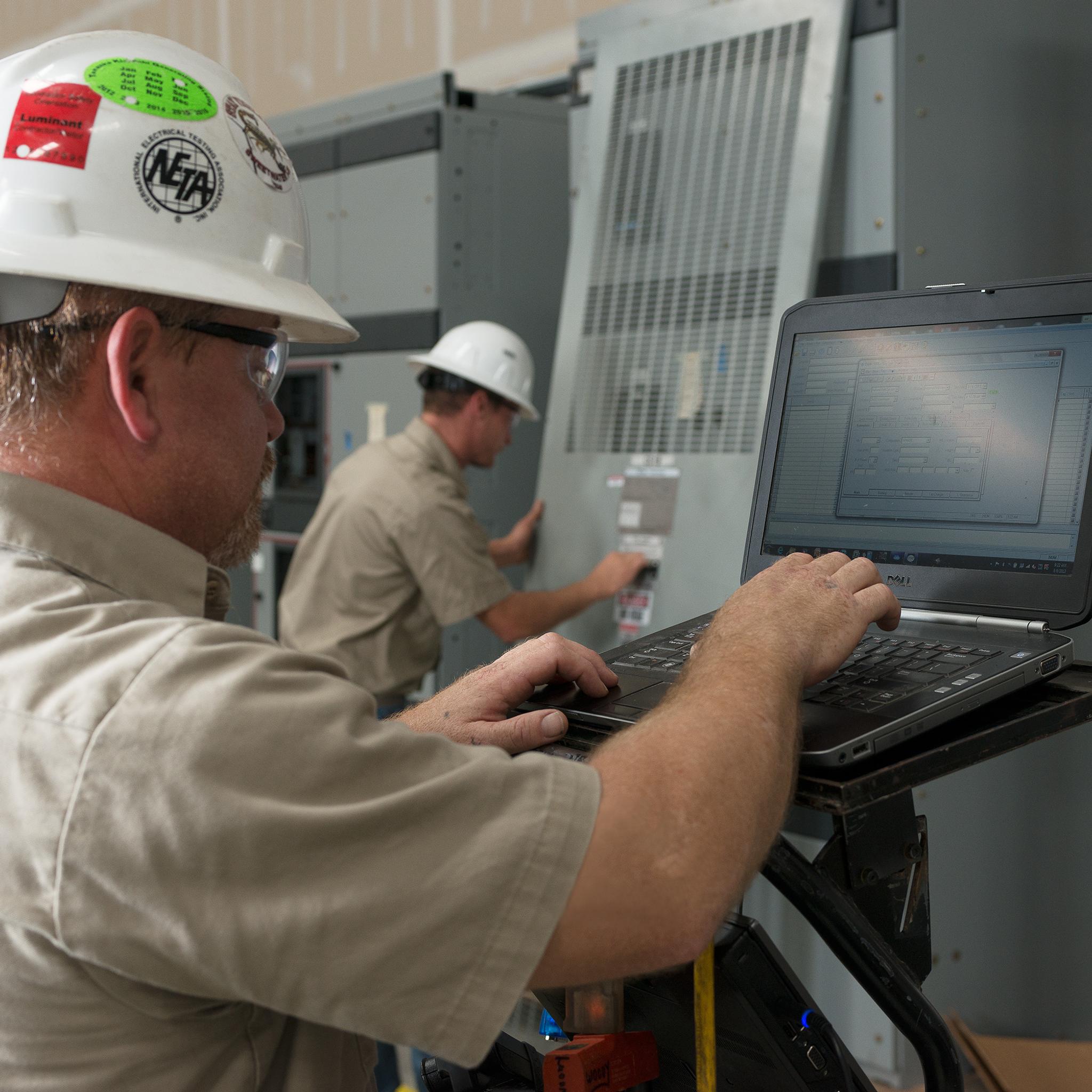 Technicians inspect industrial switchgear while one runs diagnostics on a laptop.