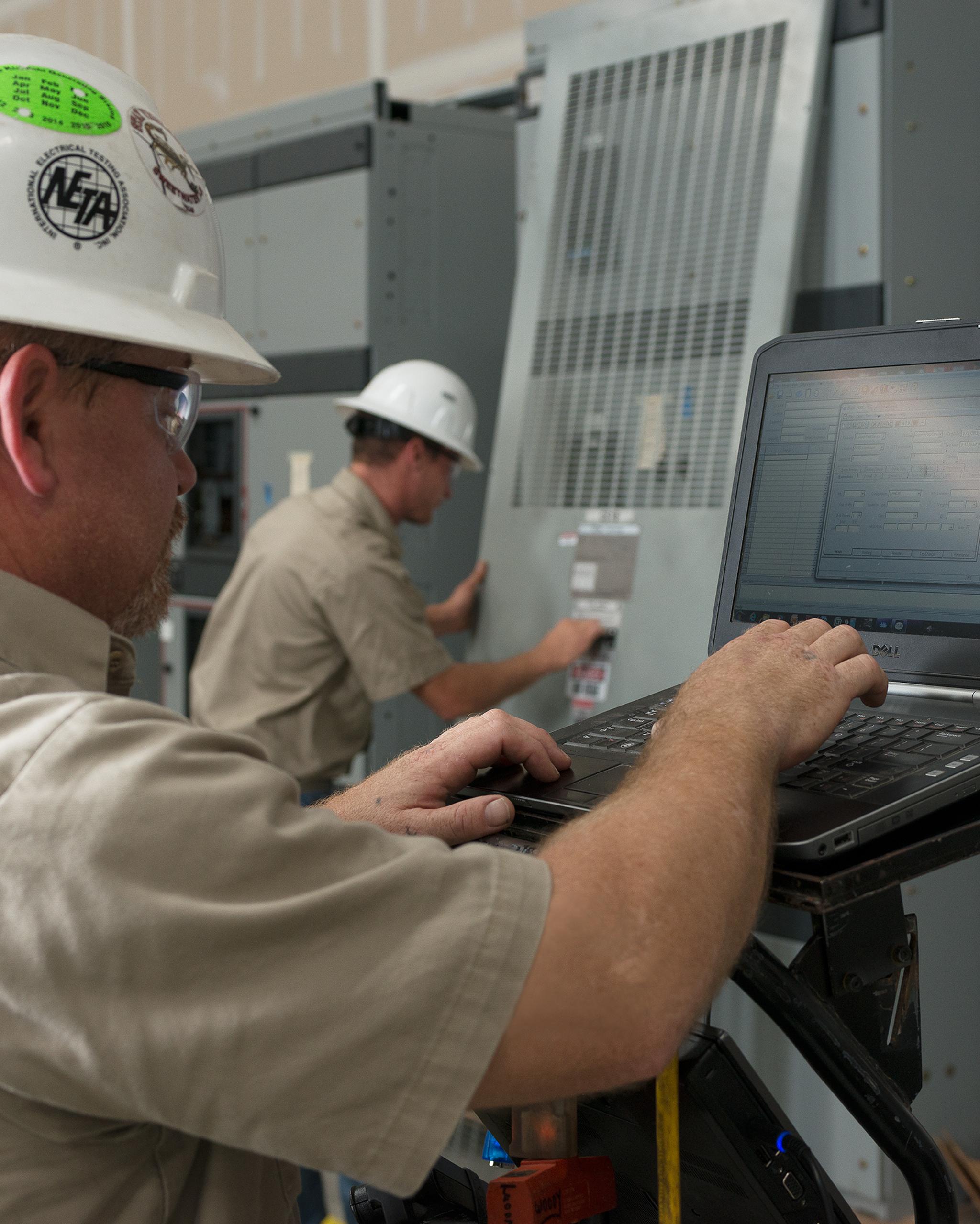 Technicians inspect industrial switchgear while one runs diagnostics on a laptop.