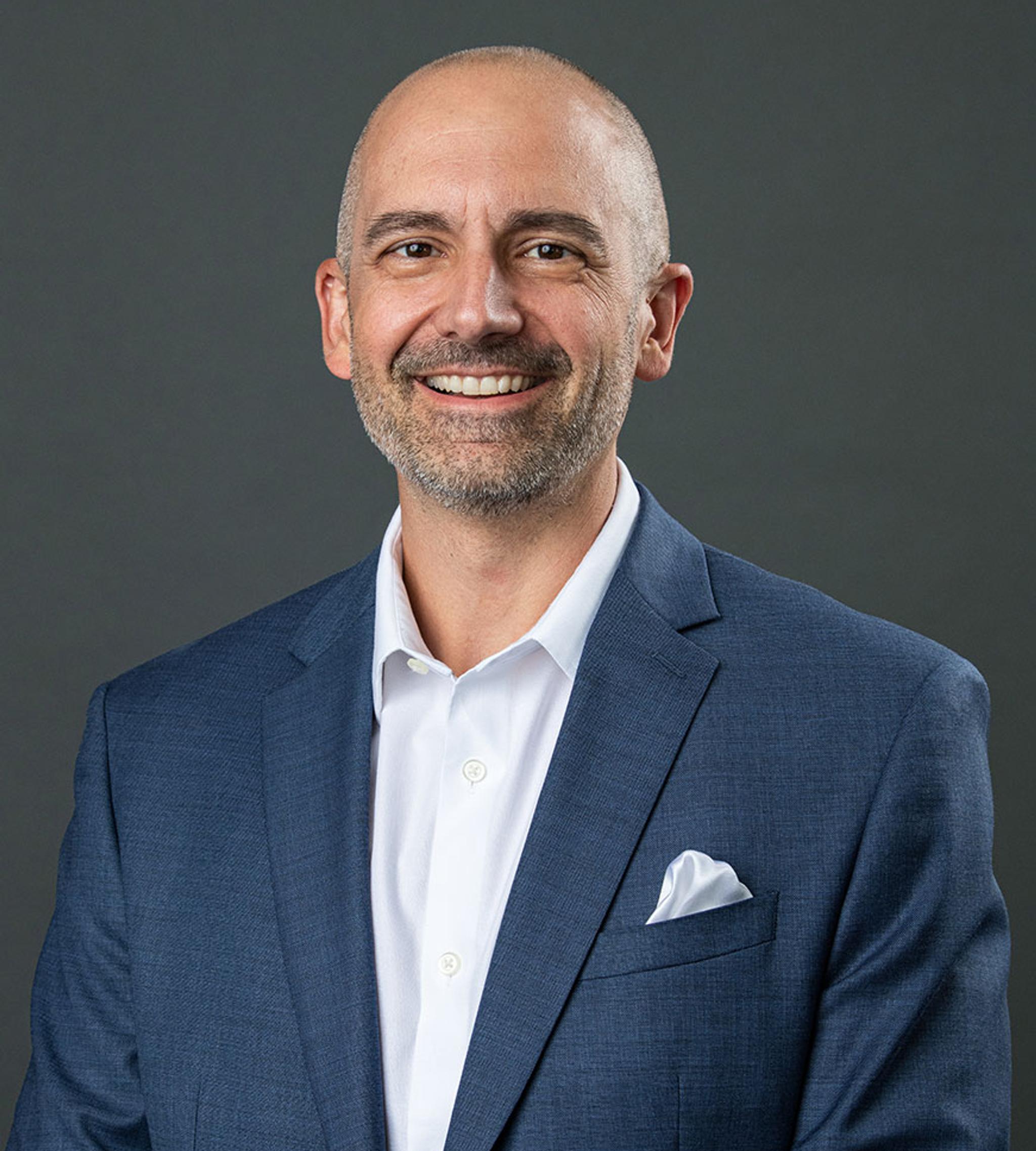 Smiling bald man with a short beard wearing a blue suit and white shirt against a gray background.