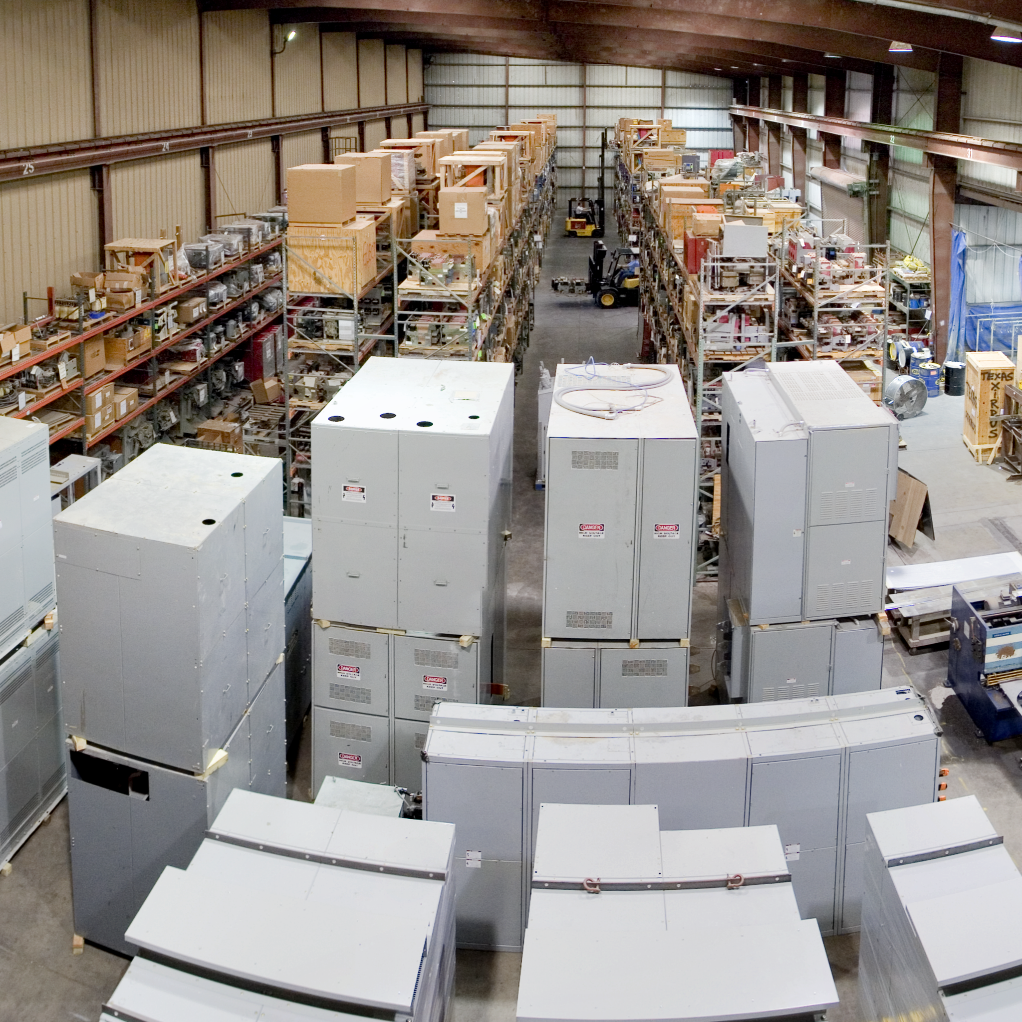 Switchgear cabinets and electrical equipment fill an industrial warehouse with stocked shelving.