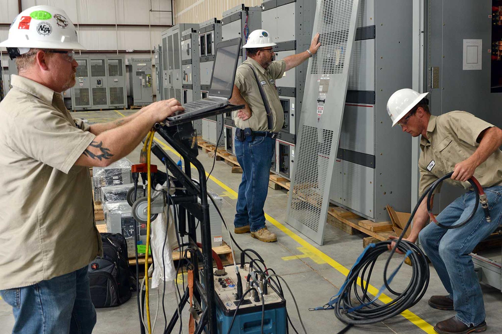 Technicians inspect and test industrial switchgear using a laptop and electrical test equipment.