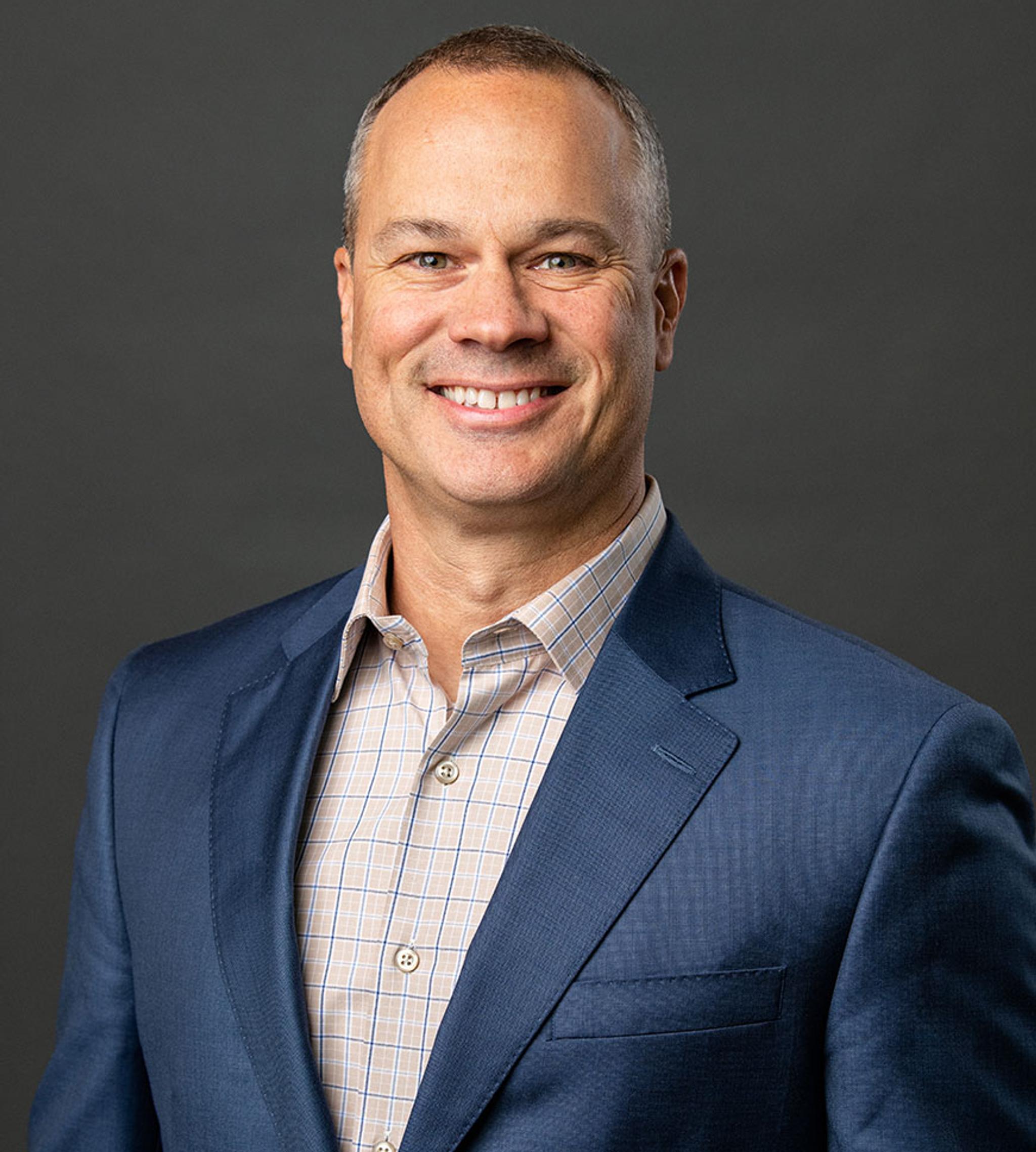 Professional headshot of a smiling man in a blue suit jacket against a dark gray background.