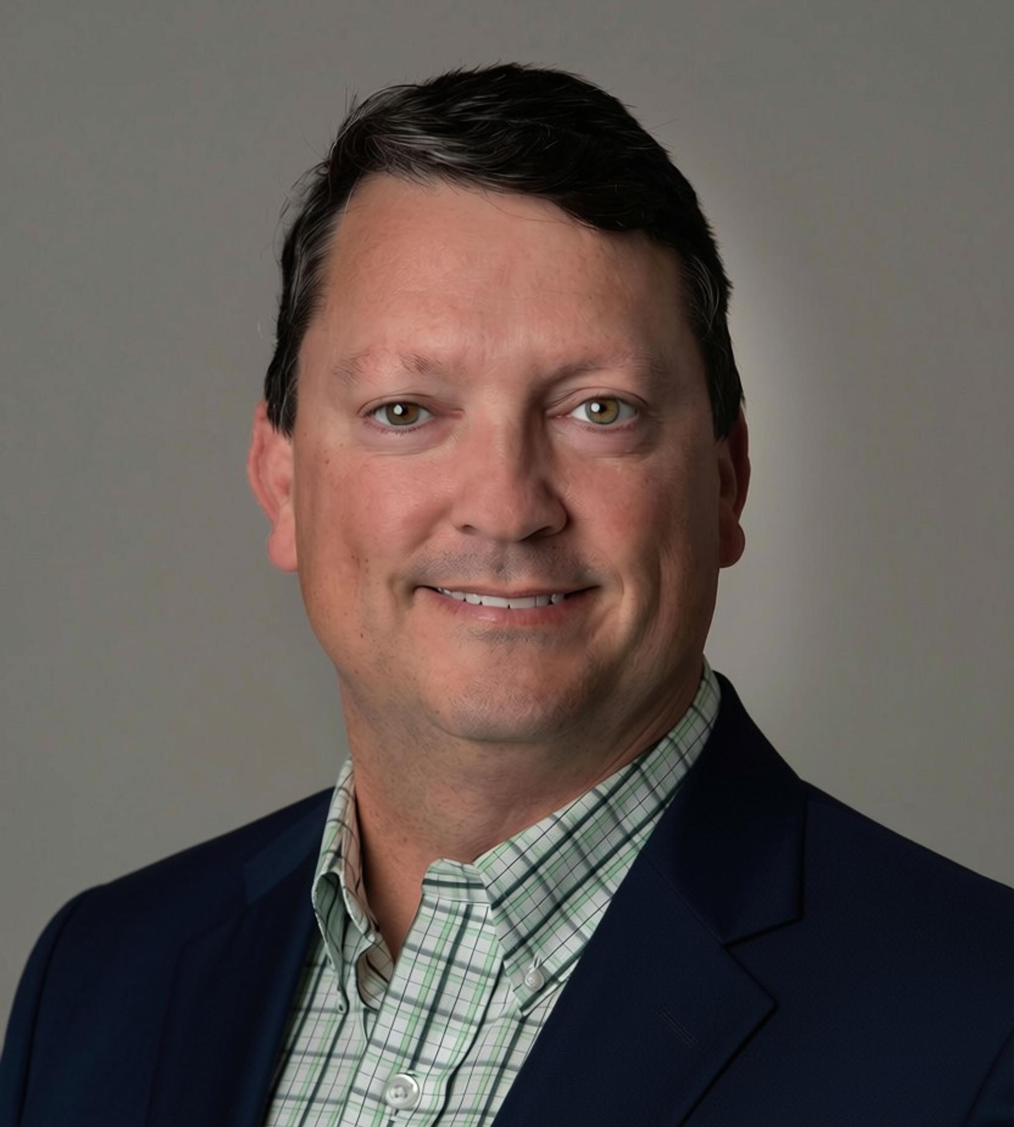 Professional headshot of a man wearing a navy blazer and checked shirt against a gray background.
