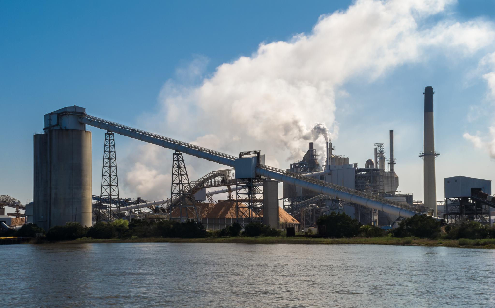 Usine industrielle avec convoyeurs, silos et cheminées rejetant de la vapeur au bord de l’eau.