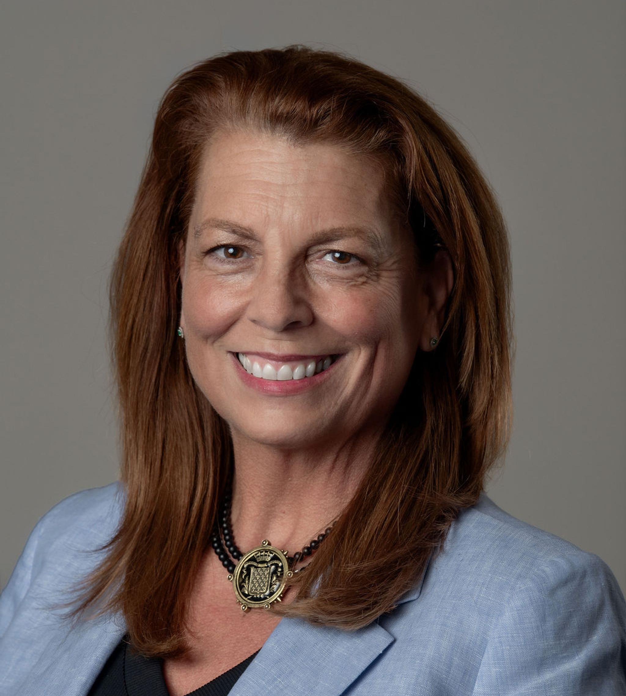 Professional headshot of a smiling woman with red hair wearing a light blue blazer and pendant necklace.