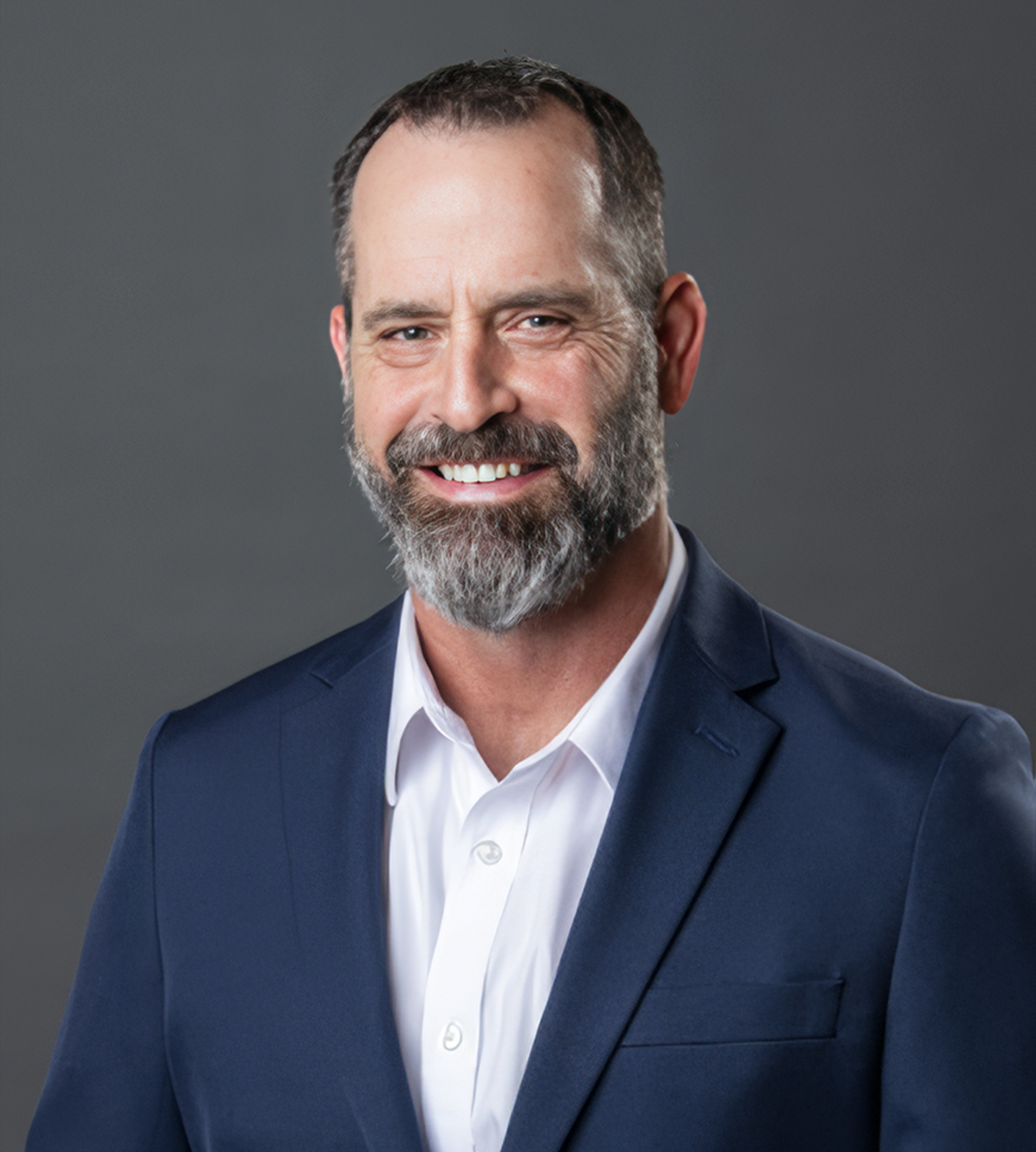 Portrait of a smiling bearded man in a navy suit and white shirt against a gray background.