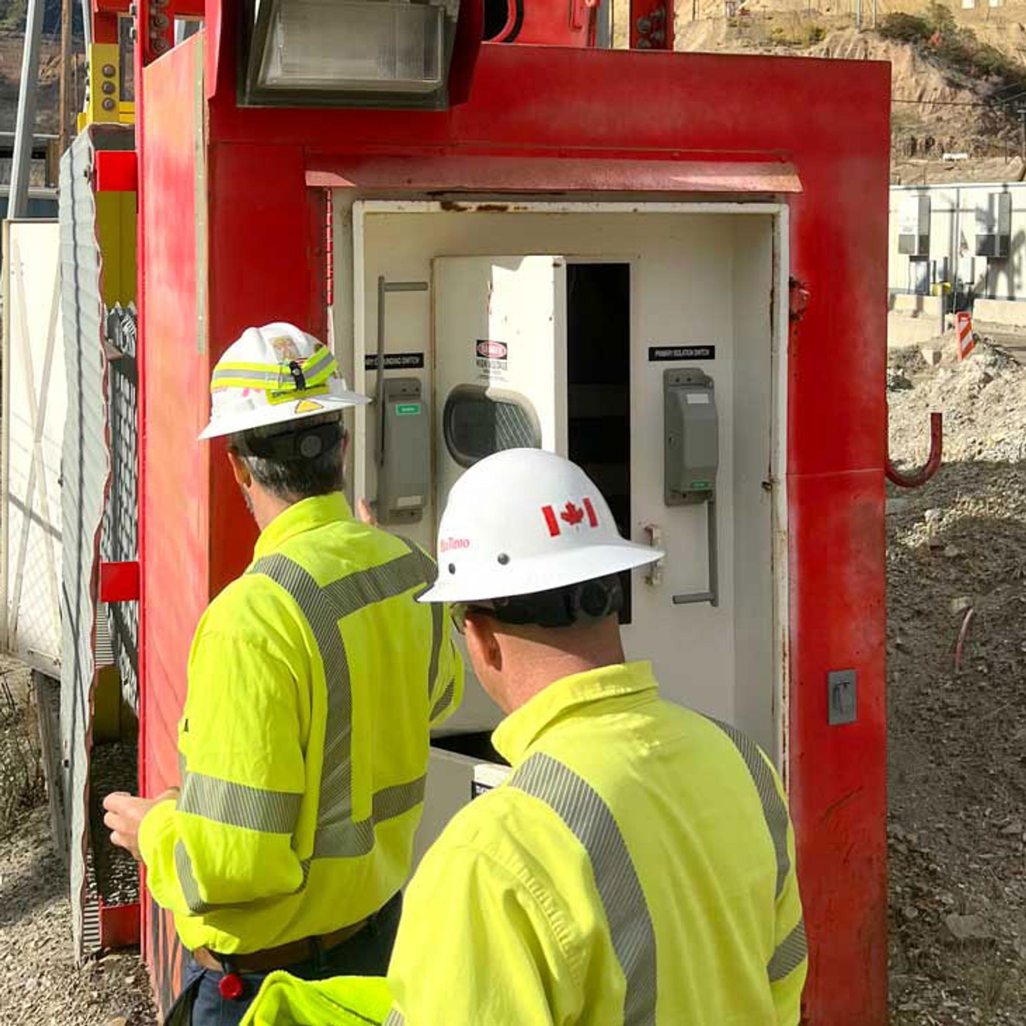 Technicians in high-visibility gear inspect an electrical switchgear cabinet at a construction site.