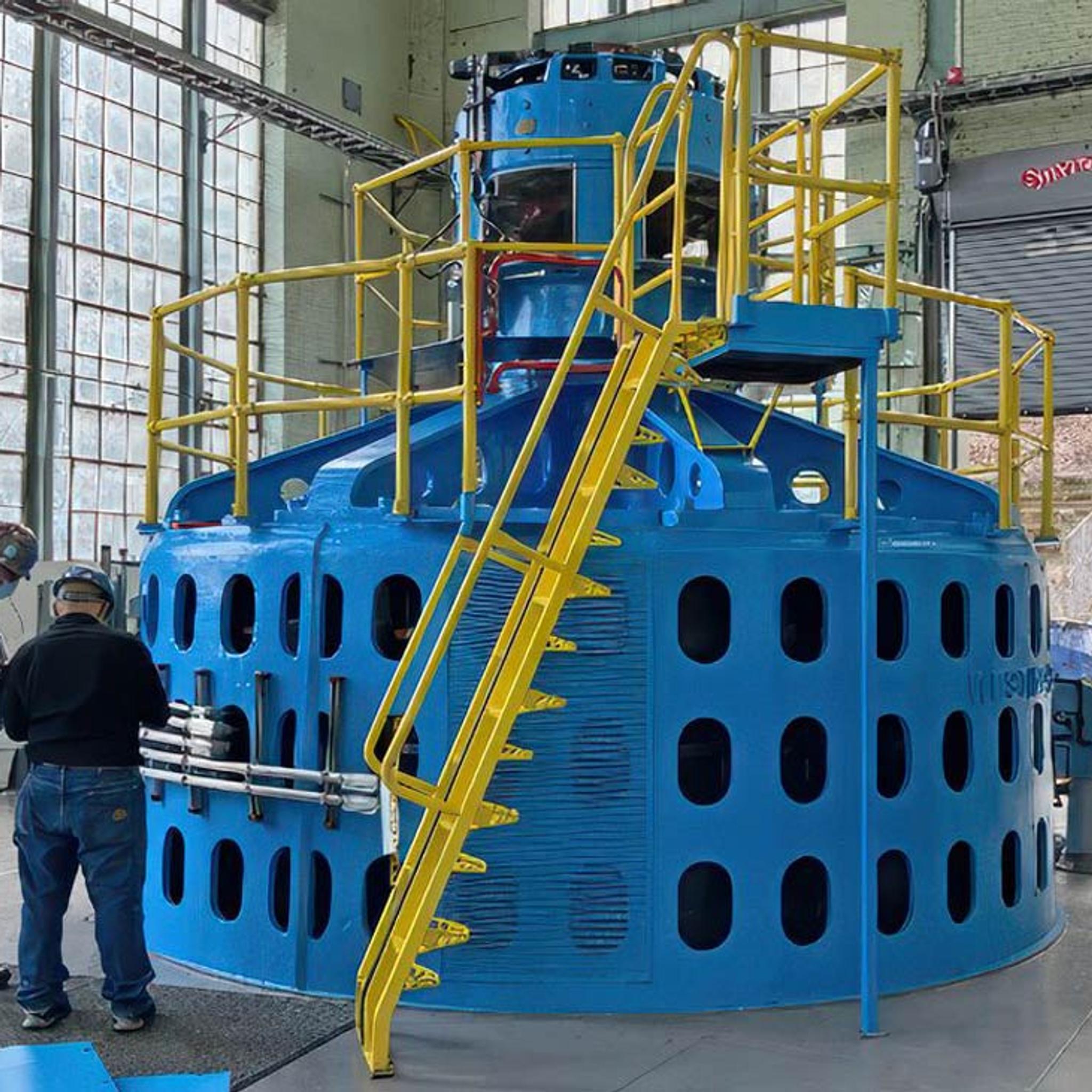 Technicians inspect a large blue generator with yellow access stairs and platforms in an industrial facility.