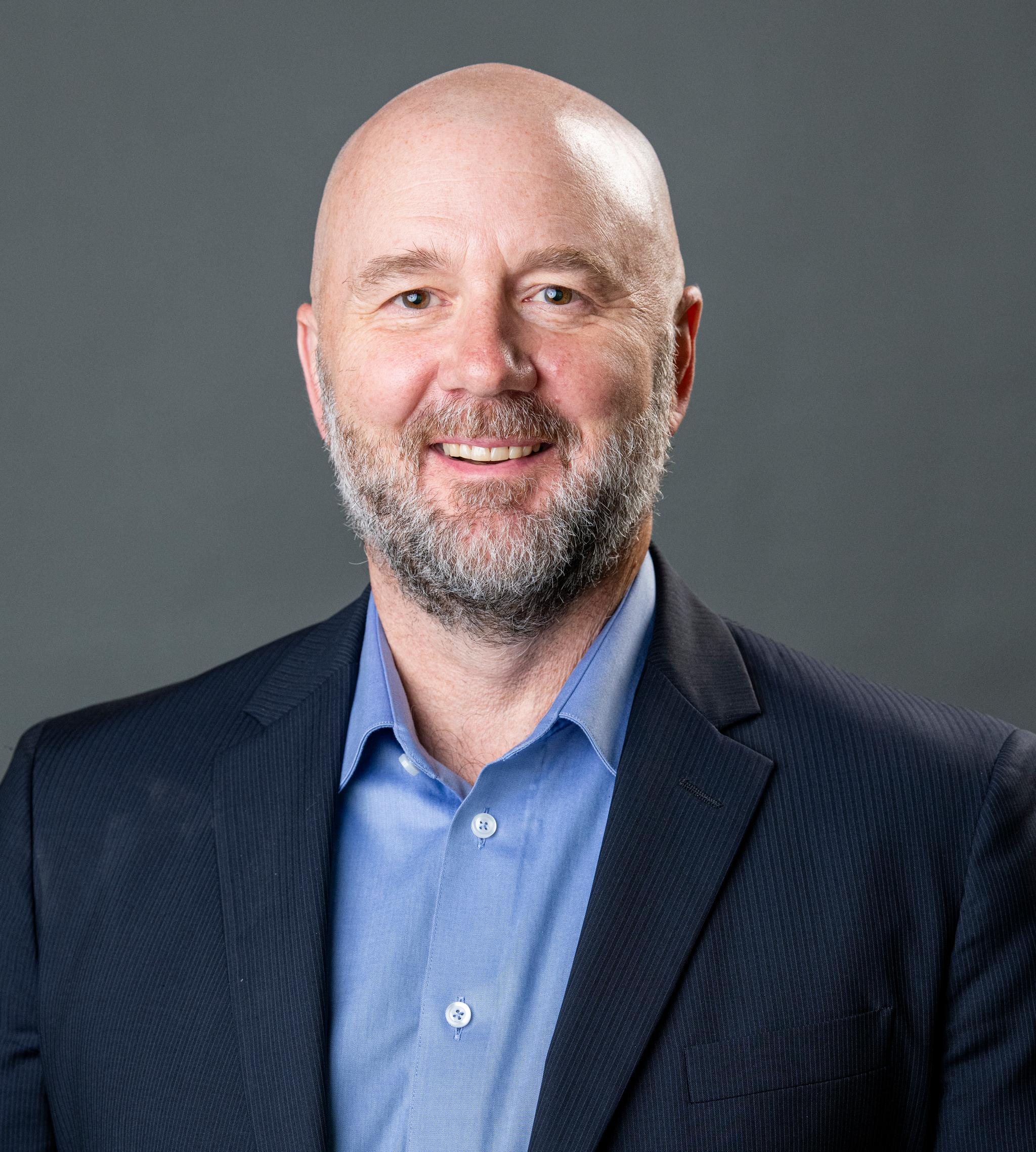 Portrait of a smiling bald man with a beard wearing a navy suit jacket and blue shirt.