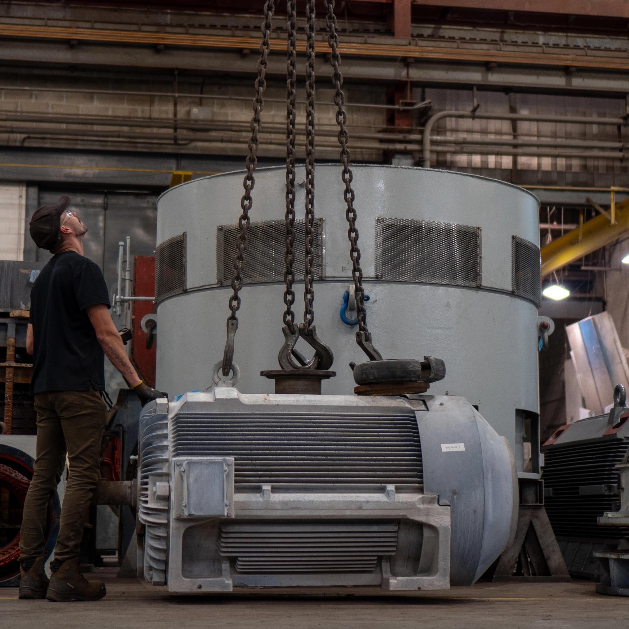 Technician guides an overhead chain hoist lifting a large industrial motor in a repair facility.