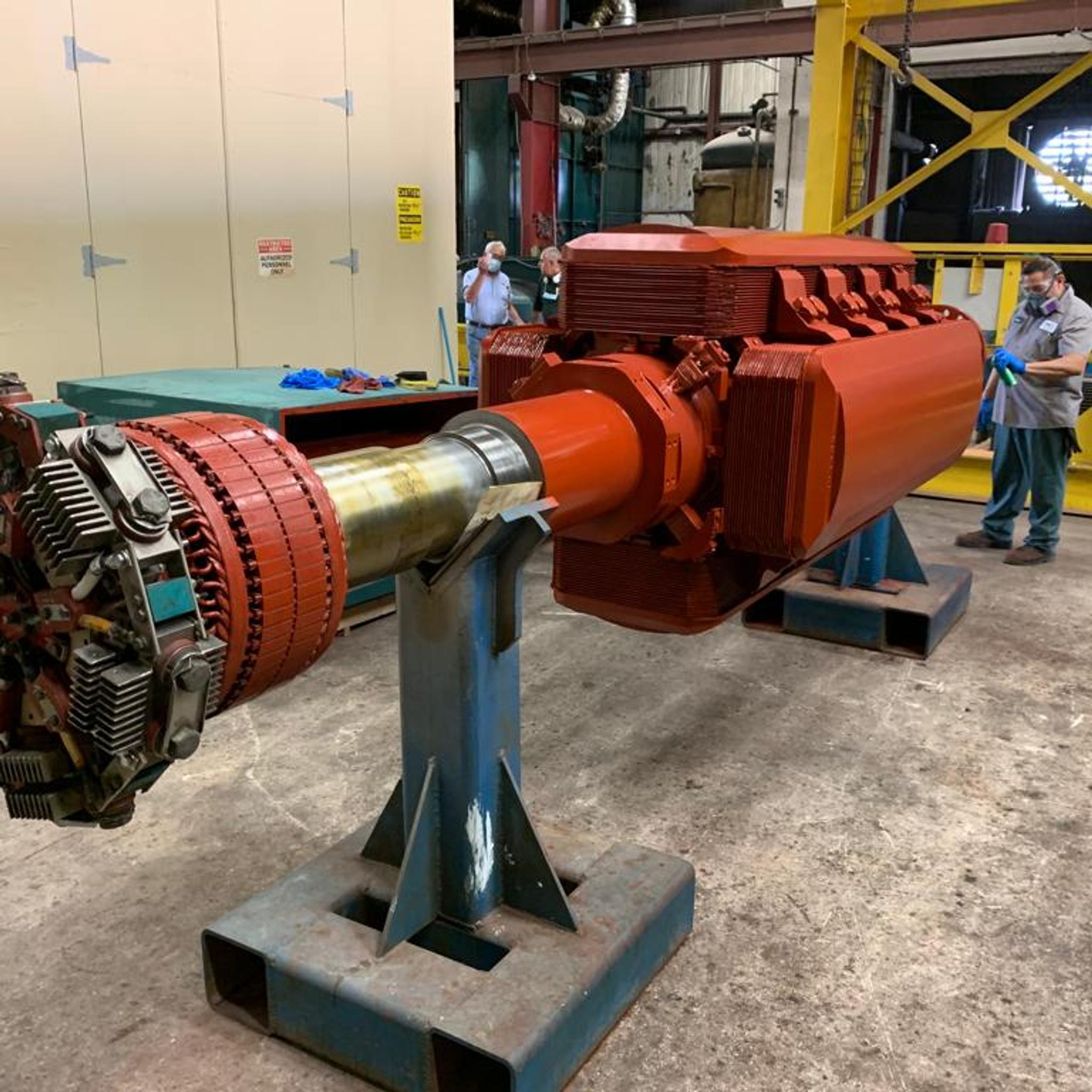 Technicians inspect a large industrial motor rotor mounted on stands in a repair facility.