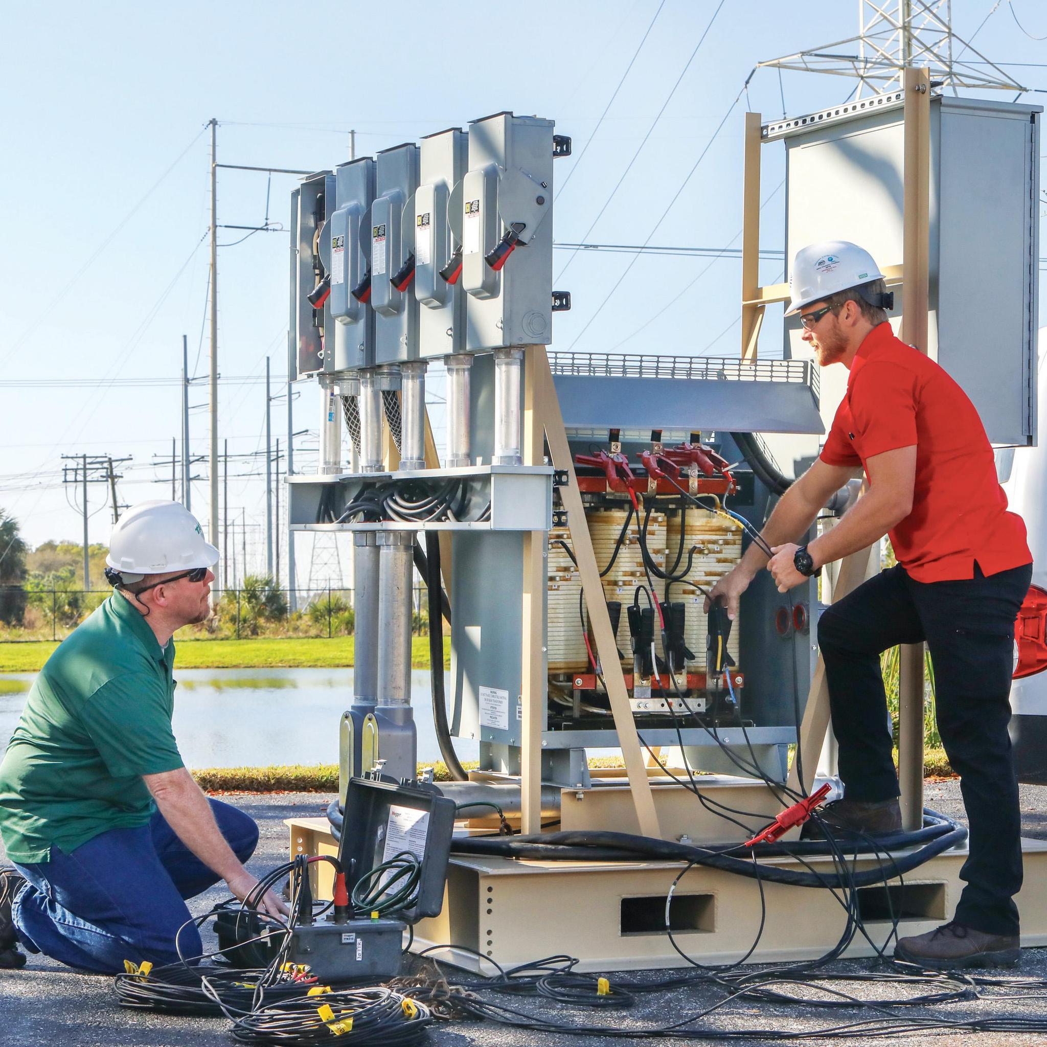 Technicians use diagnostic cables to test an outdoor electrical transformer and switchgear assembly.