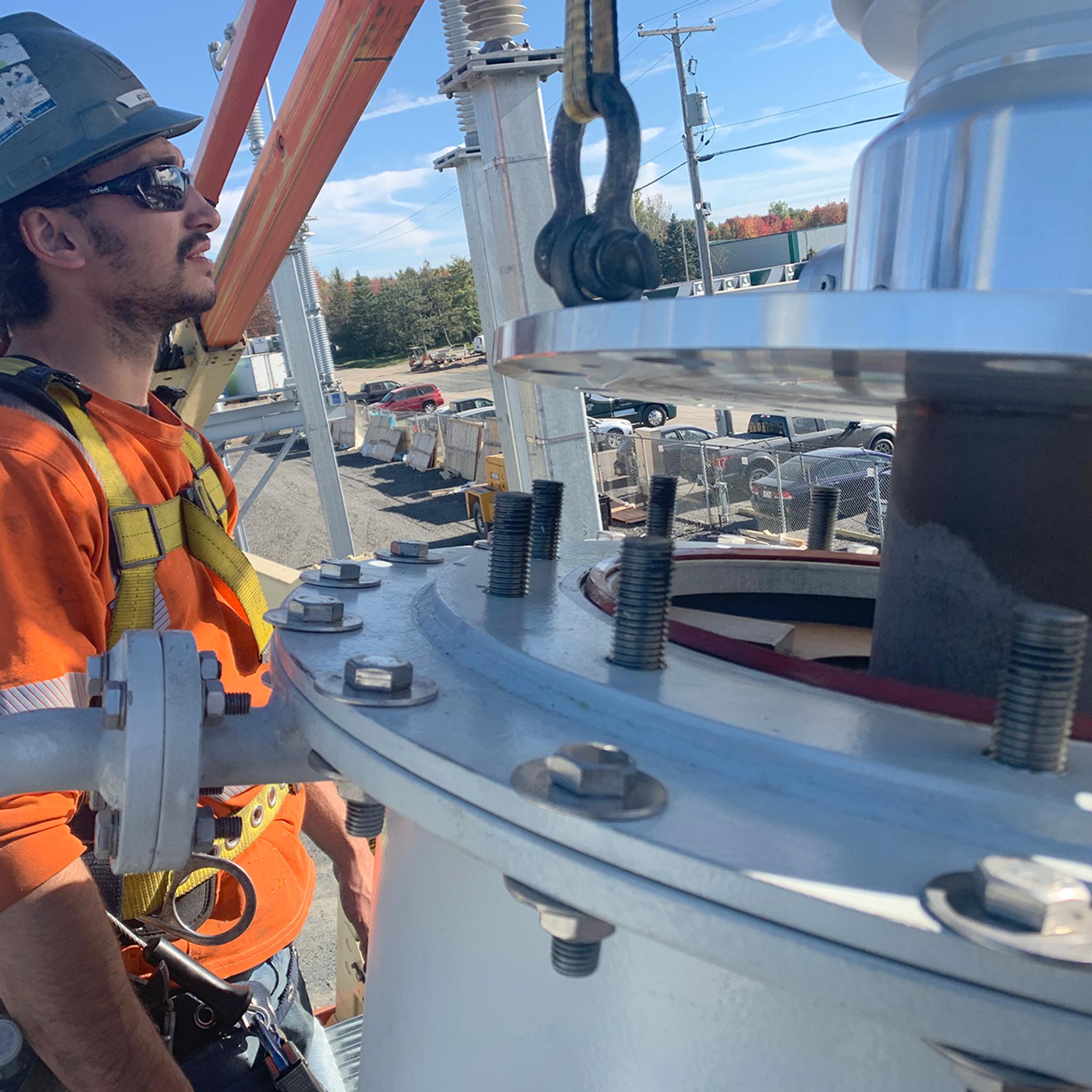 Technician guides a crane-lifted bushing into a power transformer at an electrical substation.