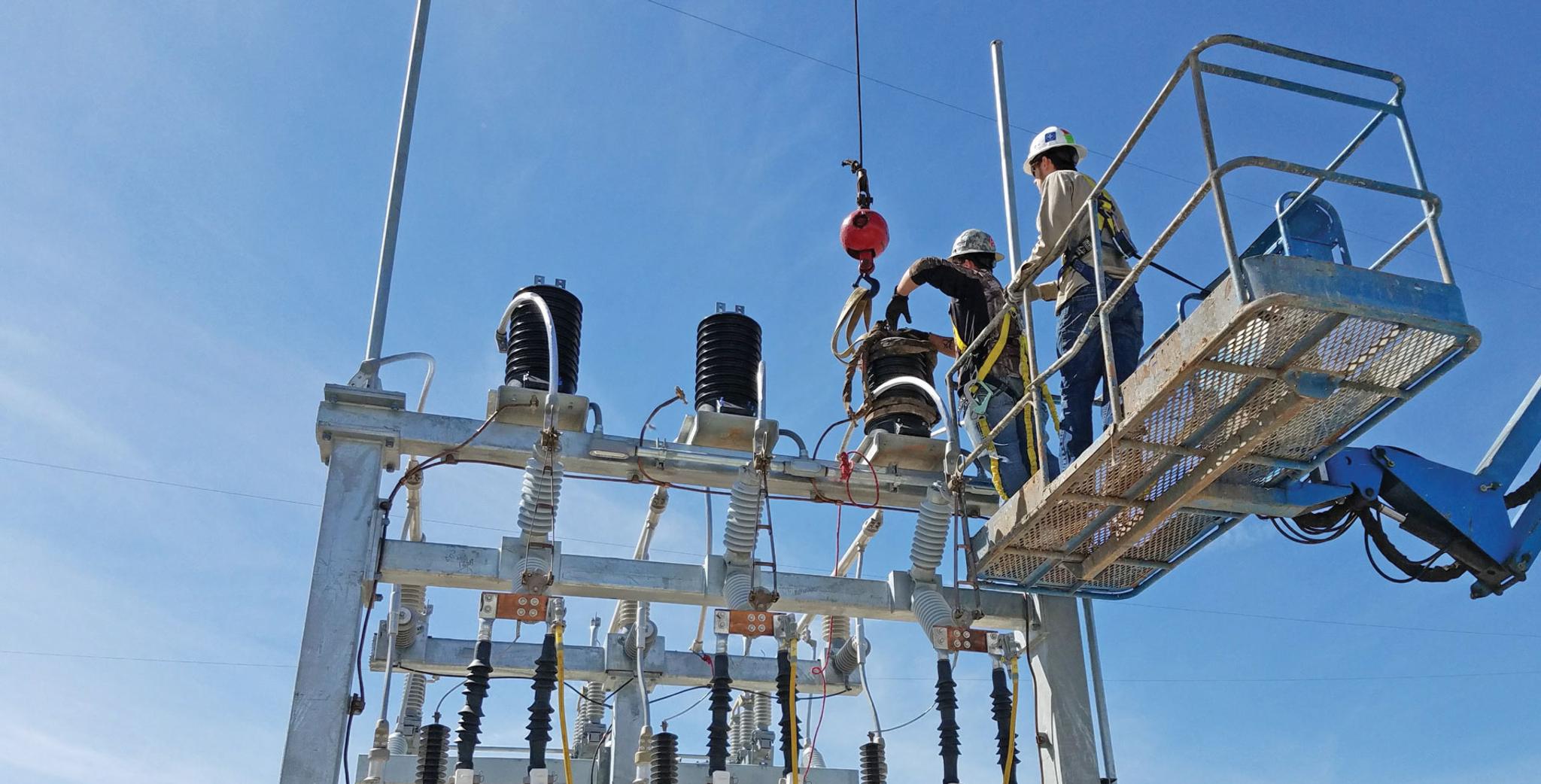 Técnicos en una plataforma elevadora instalan un aislador con una grúa en una subestación eléctrica.