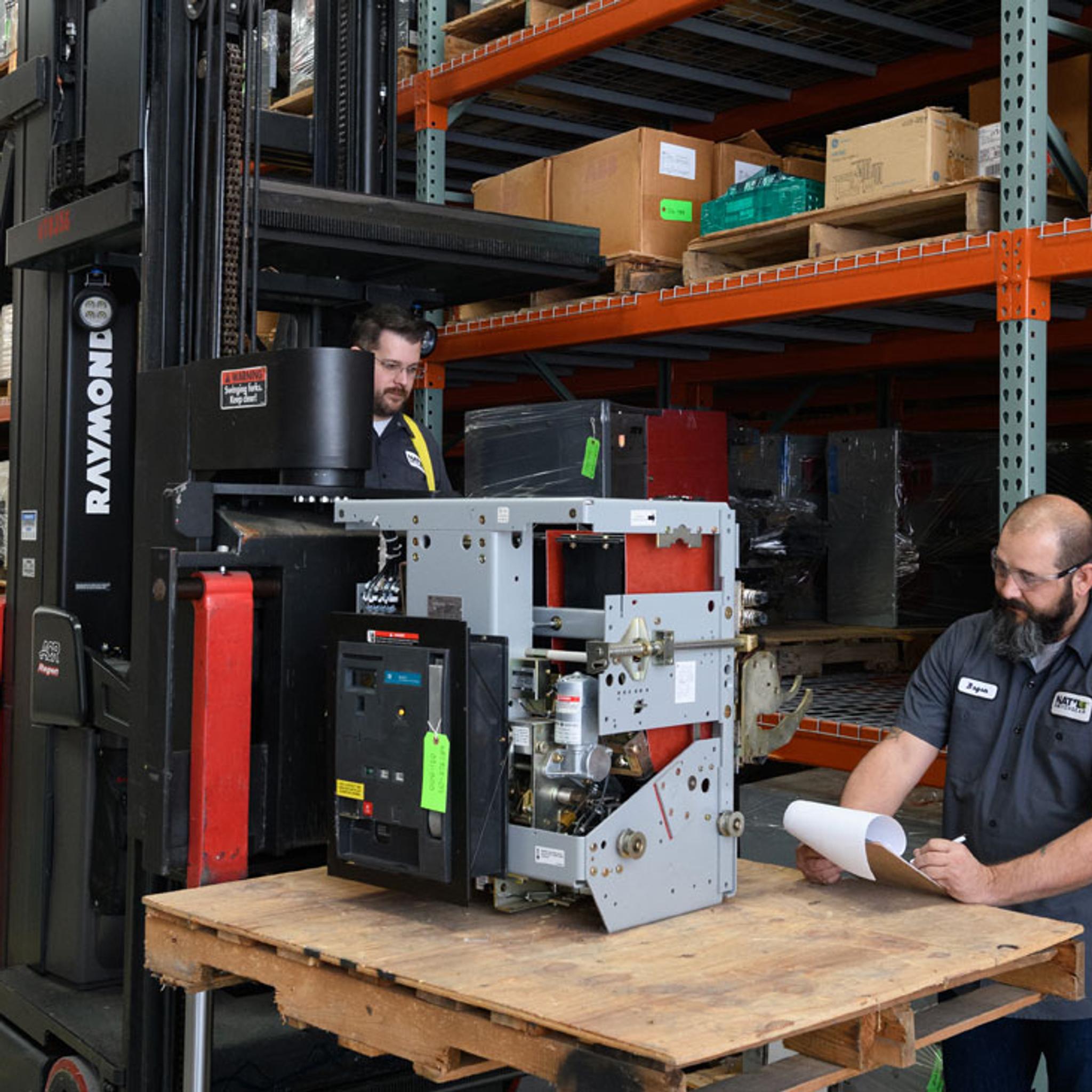 Technicians inspect industrial switchgear on a pallet beside a forklift in a warehouse.