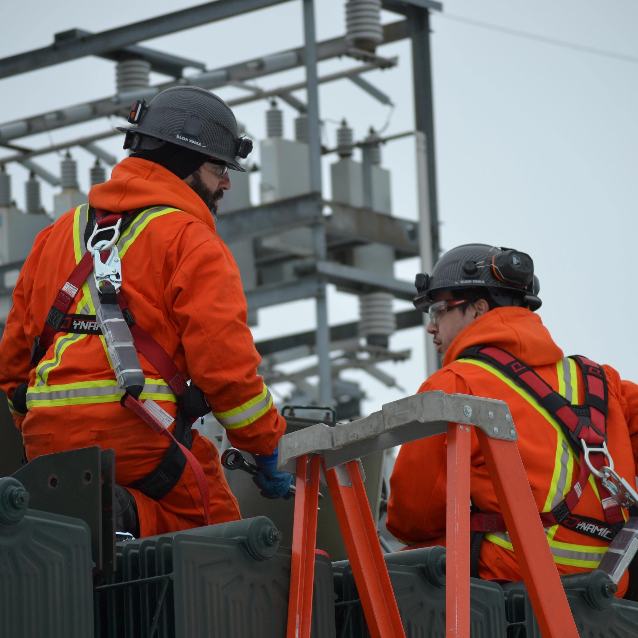 Two technicians in safety gear service electrical equipment at a high-voltage substation.
