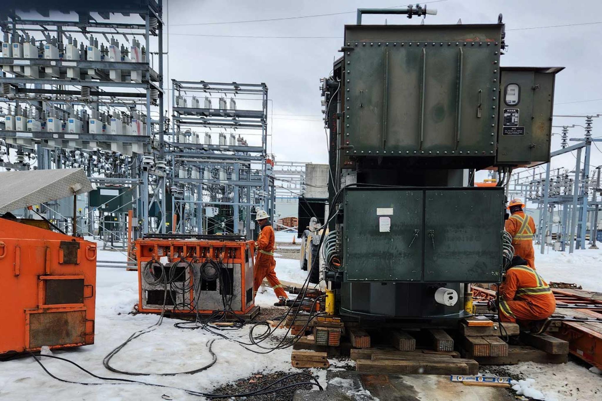 Technicians install a large power transformer at a snow-covered electrical substation.