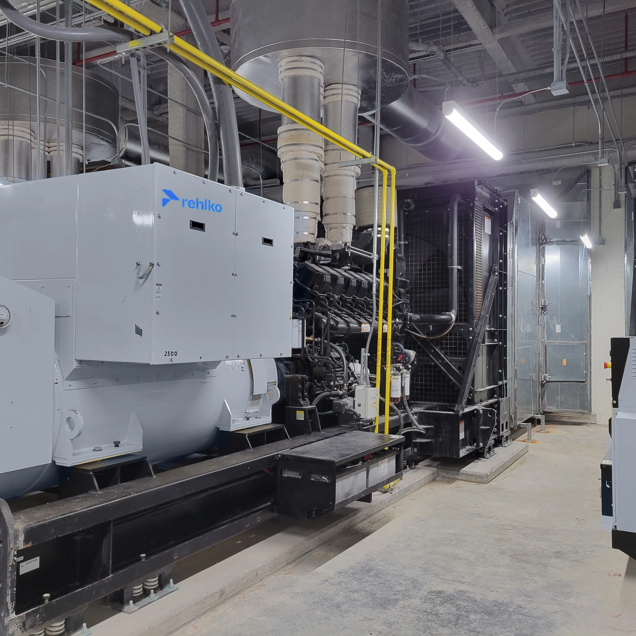 Industrial generator sets with engines, radiators and overhead exhaust ducts in a mechanical room.